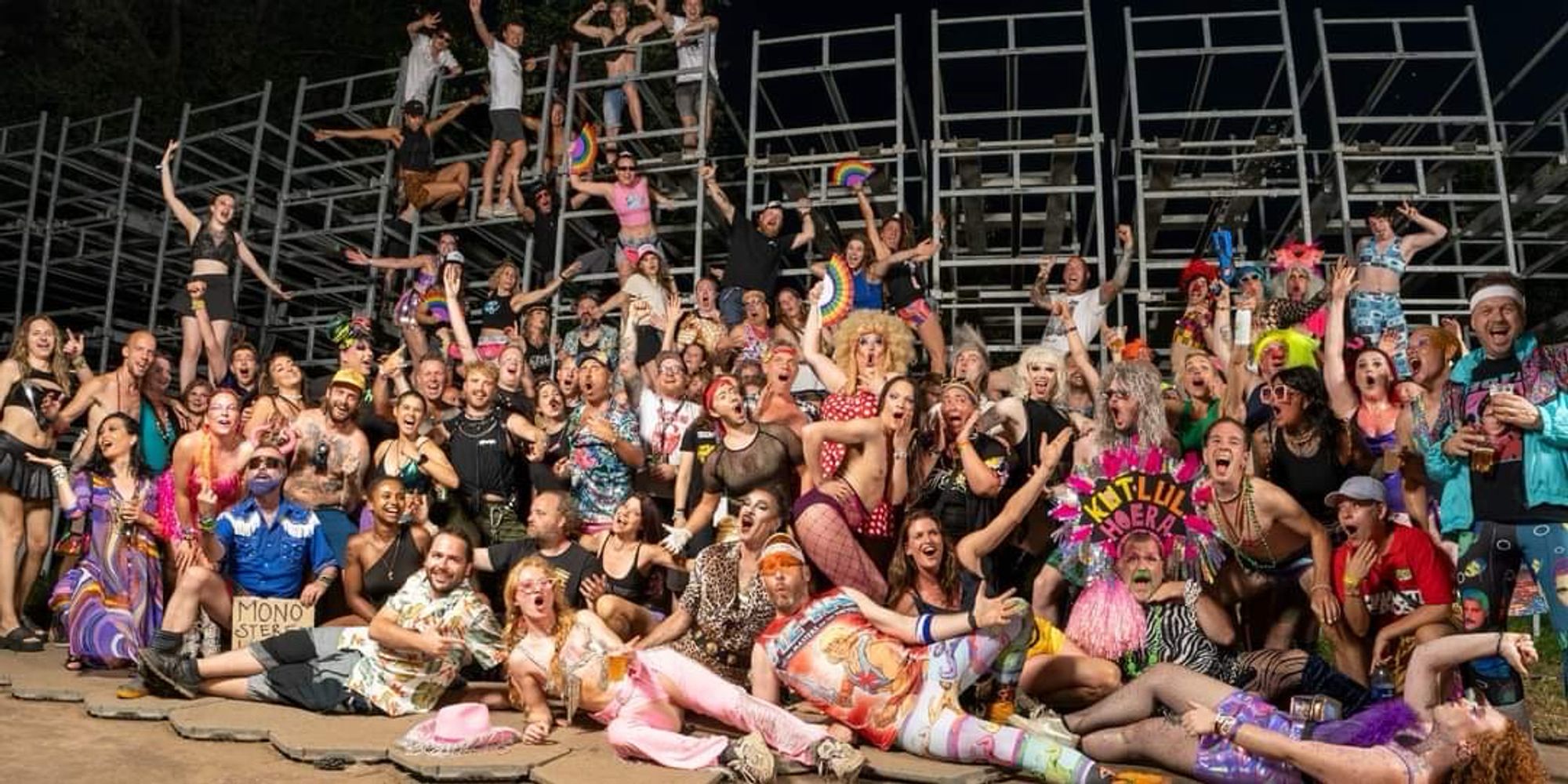 Chaotische groepsfoto met plm 150 mensen. Een paar klimmen er op het hek achter de groep. Het is een bonte verzameling van Metal- en punkliefhebbers, drags, mensen met veelkleurige kleding, iedereen lacht en joelt.