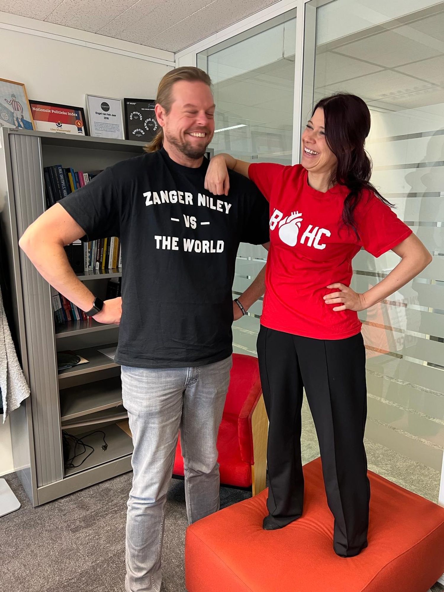 Foto van Daan in Spijkerbroek met zwart shirt en tekst ‘zanger Niley vs the world’. Ik sta naast hem op een rode kussentafel en draag een rood shirt van de Jeugdbende. Achter ons een boekenkast en een raam naar de gang.