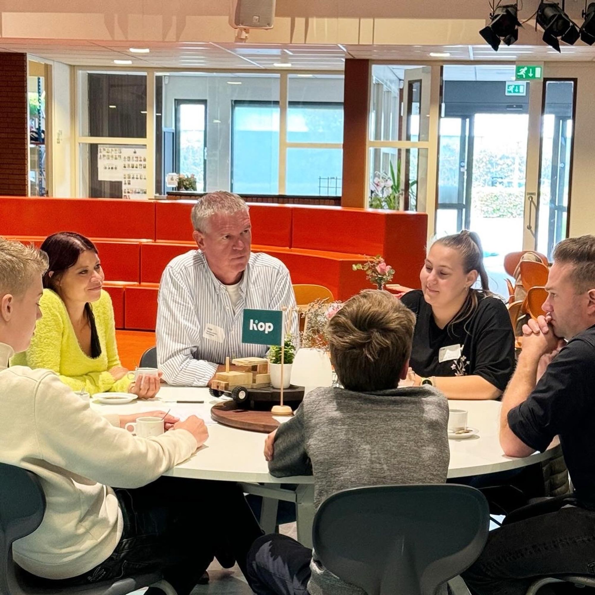 Foto van een witte tafel met daaraan 6 mensen (7e is net uit beeld) waarvan twee leerlingen, begeleider, twee personen uit het bedrijfsleven en ik. We zijn met elkaar in gesprek, op de tafel staan koffiekopjes en wat onduidelijke houten attributen in het midden. Op de achtergrond rode blokken.