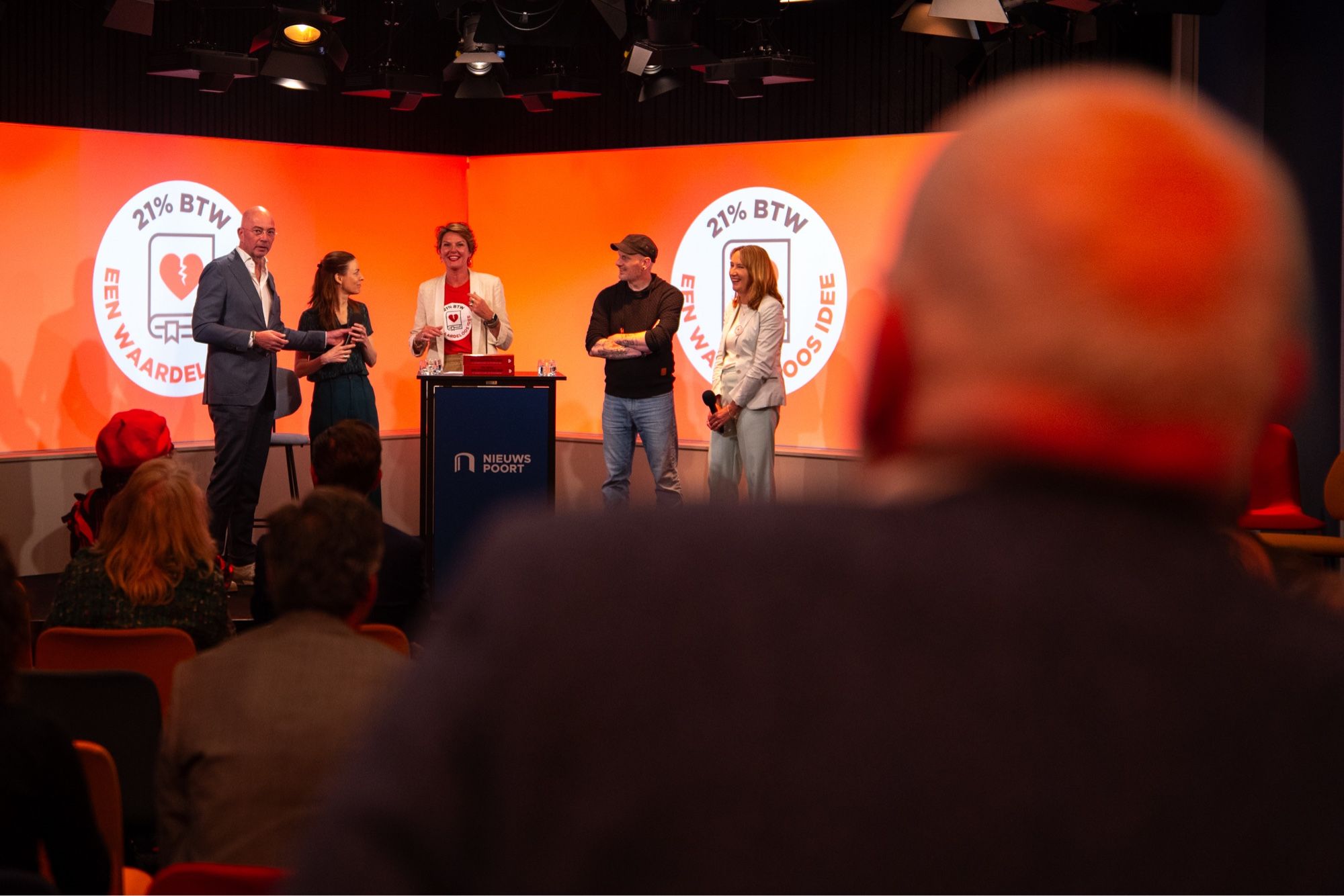 Ik zit achter Frans Timmermans in een zaaltje met uitzicht op het podium waar diverse mensen aan het praten zijn. Op de muur weer het logo.