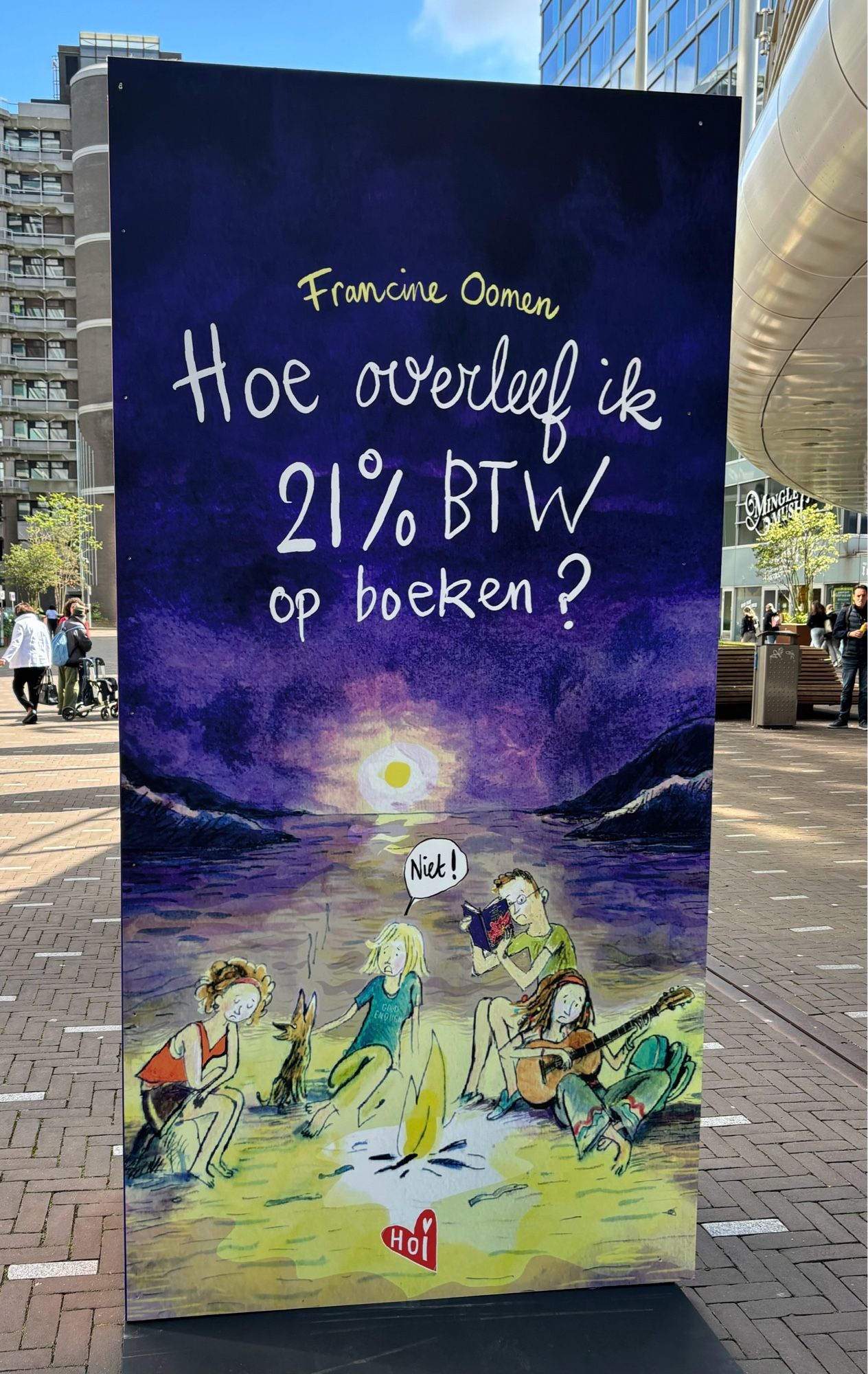 Foto van een ruim 2 m hoog staand boek van Francine Oomen, met de titel “Hoe overleef ik 21% BTW op boeken?” Met een tekening daarover van een aantal jongeren rondom een kampvuur, voor een getekende zee met ondergaande zon en zwartblauwe lucht.