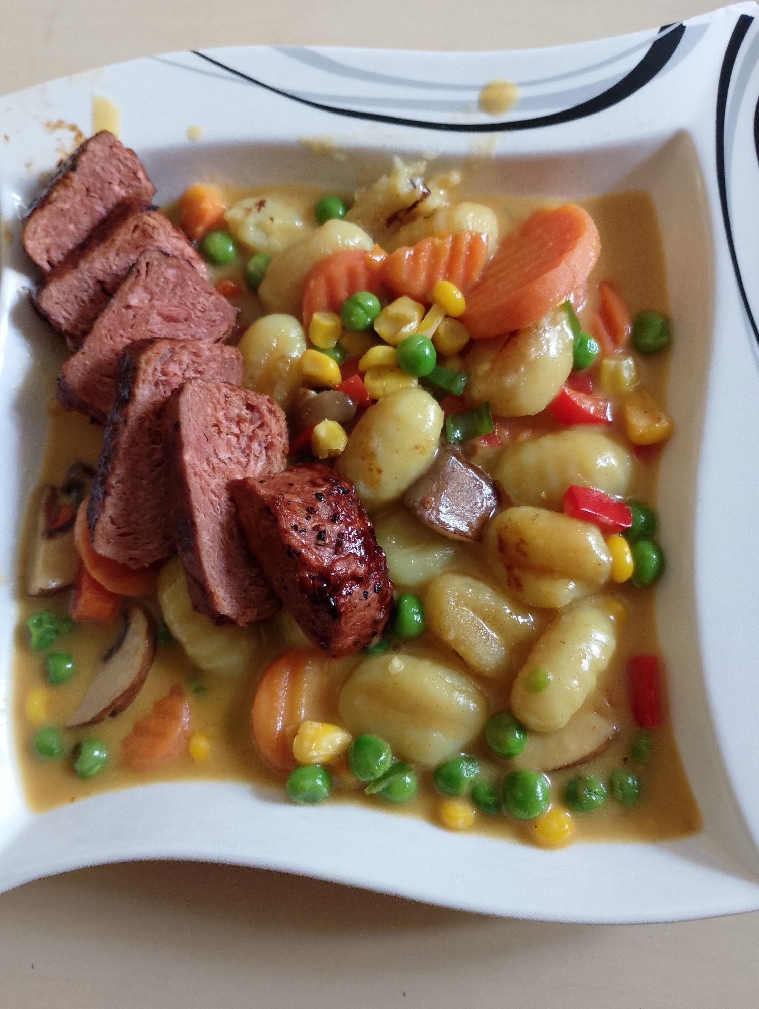 Teller mit Gnocchi, Möhren, Mais, Paprika und Erbsen in einer hellen Soße, dazu in Scheiben geschnittenes Steak