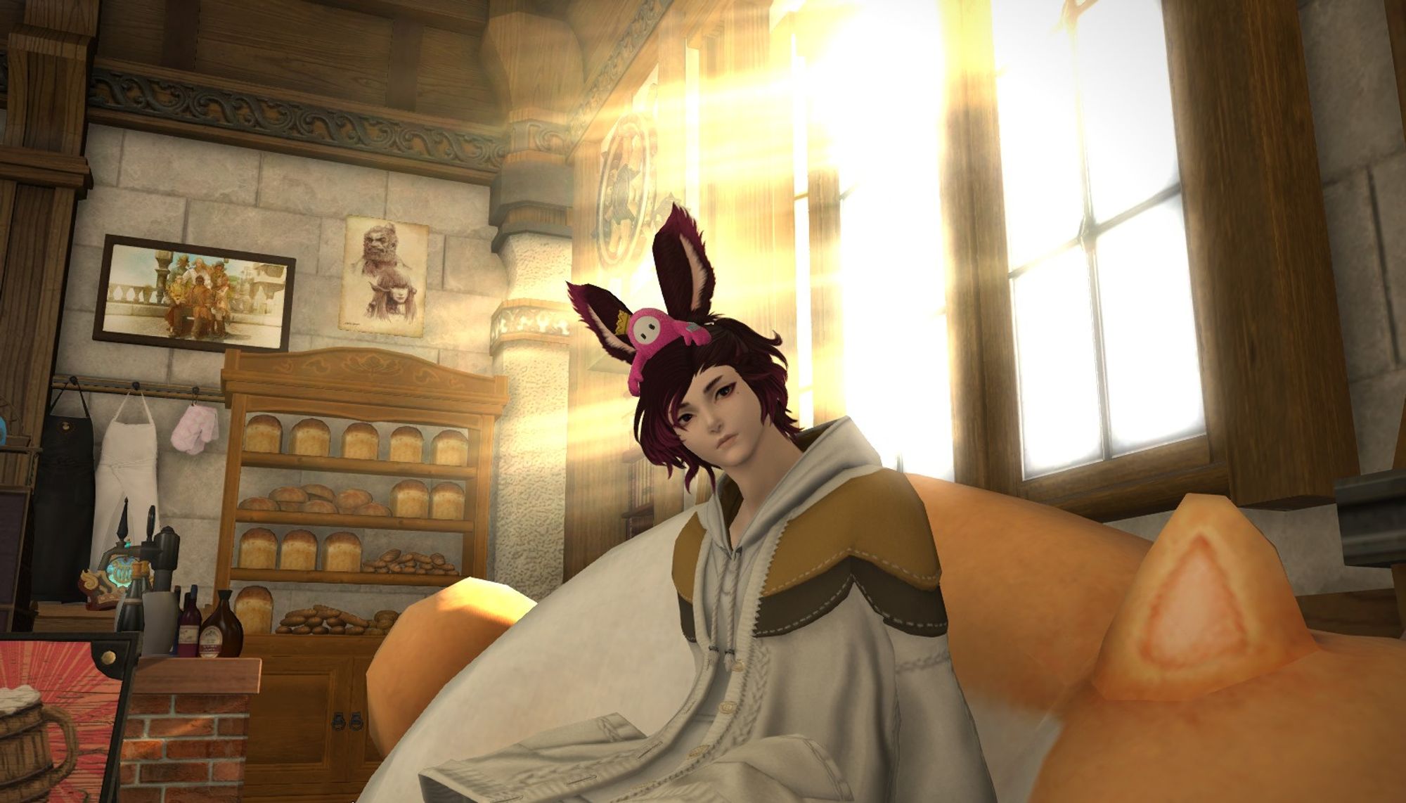 An original video game character from the game Final Fantasy XIV. The character is a male viera. He is wearing a white hoodie with light brown and dark brown on the shoulders, made to mimic the patterns of a cat from the video game. He has medium length brown hair with purple tips. His bunny ears are dark brown as well with purple tips. He has a small pink bean character on his head as a hat from the video game Fall Guys. He is sitting on a couch made to look like a cat from FFXIV in a cafe. He is in front of two windows with bright sunlight shining through them. In the background there is a shelf with various types of bread on it. On the wall there is a framed photo, a poster, and some hooks with aprons hanging on them. There is a brick counter with a few bottles on it and a red standing sign advertising drinks in front of the counter.