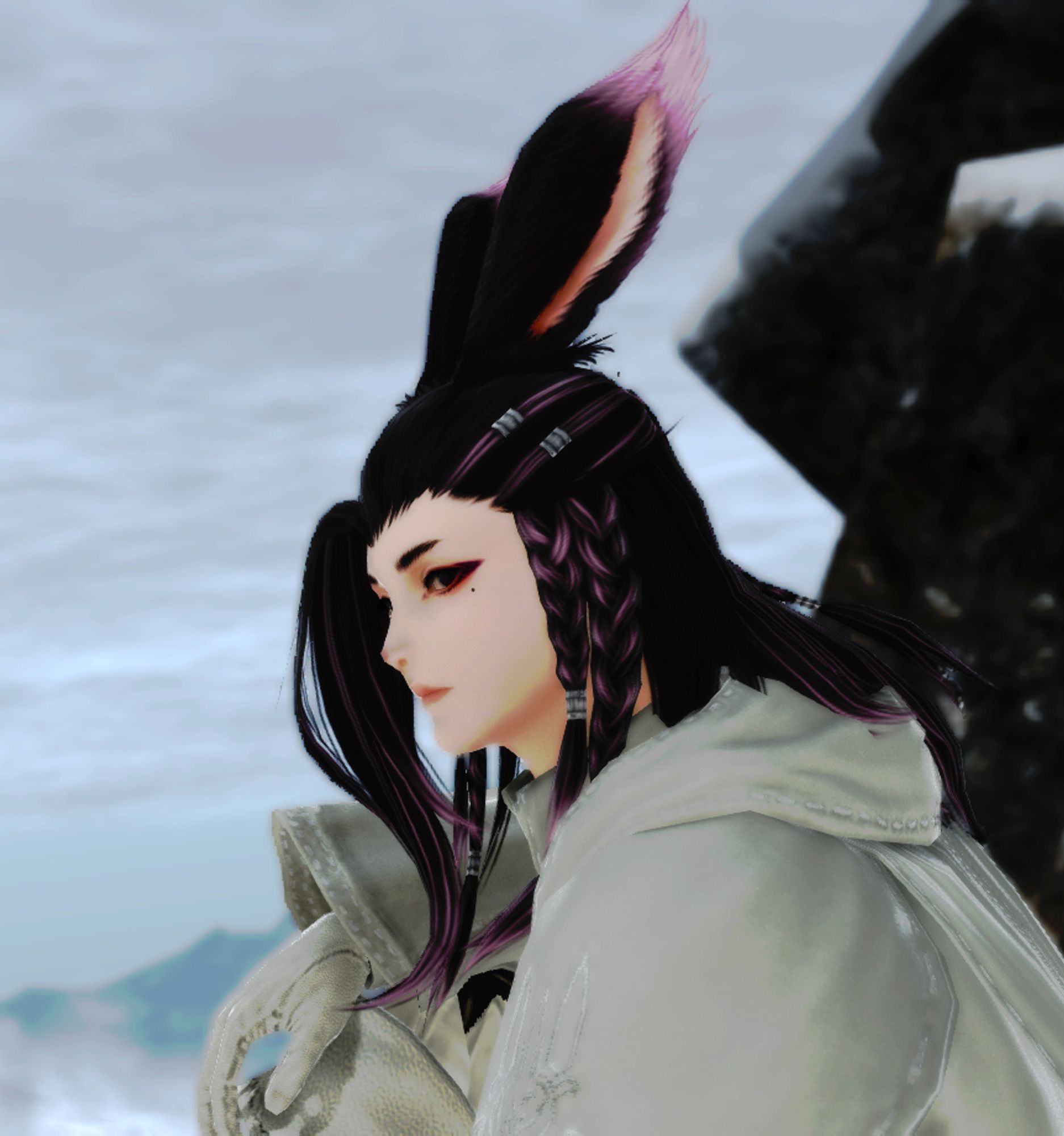 Image of an original character from the video game Final Fantasy XIV. The character is a male viera, or bunny person. He has long dark brown hair with light purple to white ends, two braids on the left side, and two braids on the right side. He has long bunny ears that stand upright with purple to white tips. He is wearing an all-white hooded top with white gloves and is looking to the left in the photo. He is sitting up on a snowy mountain with the mountain visible in the background of the photo. The environment is gray and foggy.