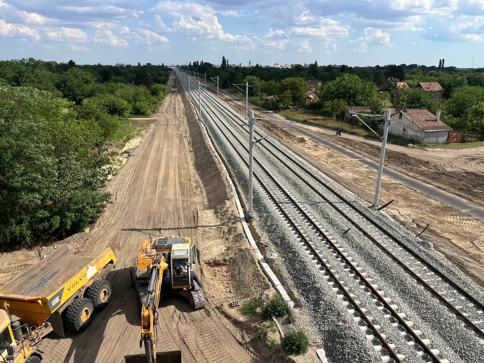 A wide view shows a railway construction site. On the left, heavy machinery, including an excavator and a dump truck, are parked on a dirt road. On the right, freshly laid train tracks extend into the distance, flanked by green trees and scattered houses under a partly cloudy sky.
