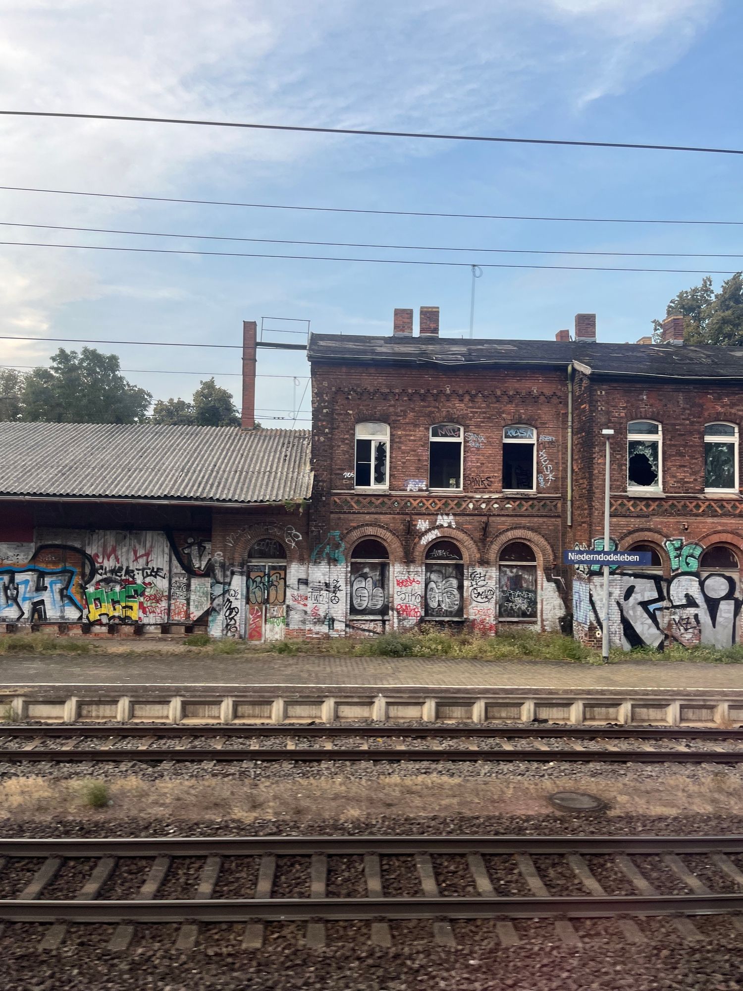 Station Niederndodeleben. A two storey brick building to the right, a single storey kind of goods shed to the left. Broken windows. Covered in graffiti. Tracks and platform in front of the building 