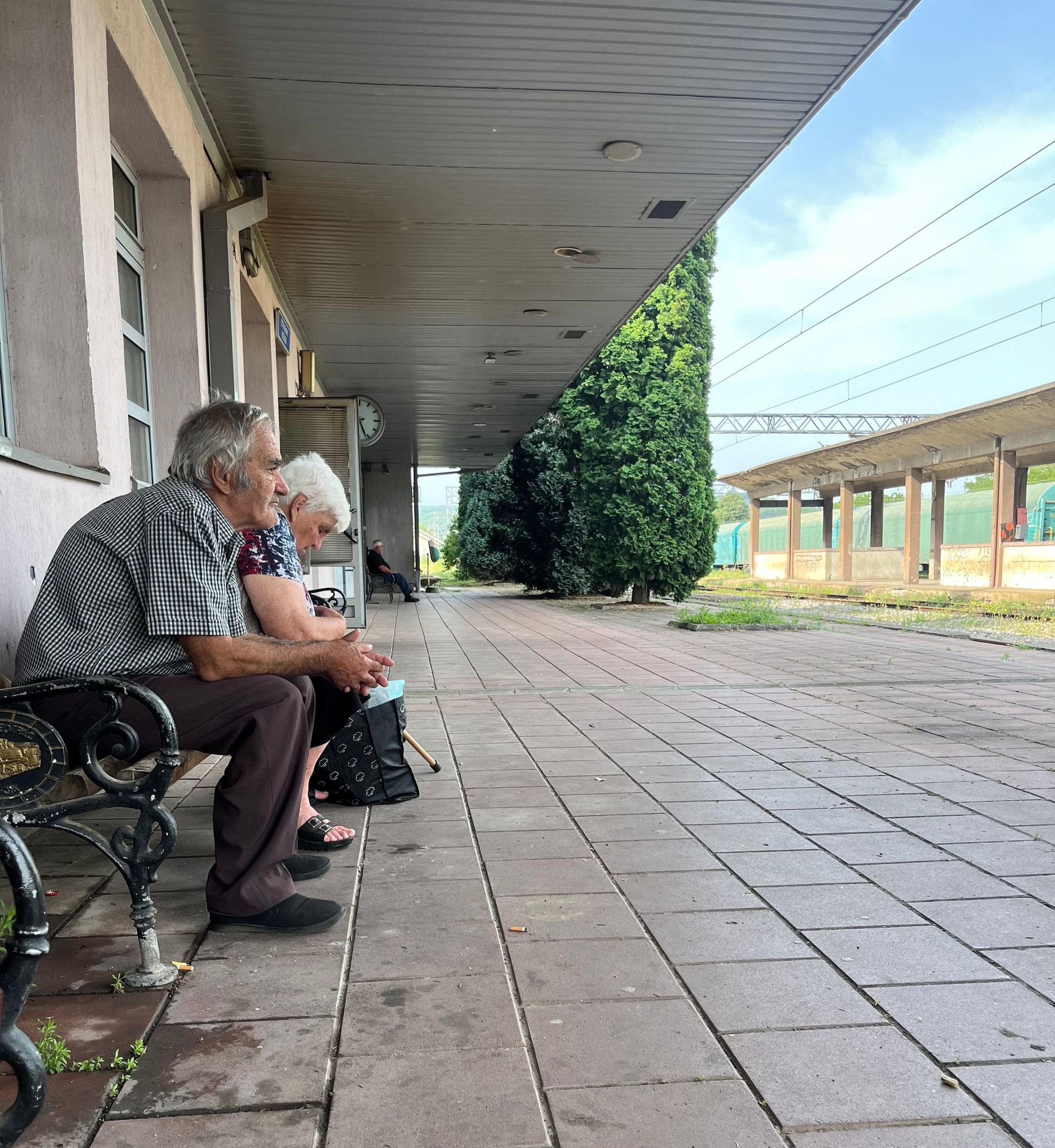 An elderly man and woman sit on a metal bench under a covered platform at a train station, gazing into the distance. The platform is empty and has a tall trees on it in the distance. The sky is clear, and train tracks run parallel to the platform, leading to a partially visible underpass.
