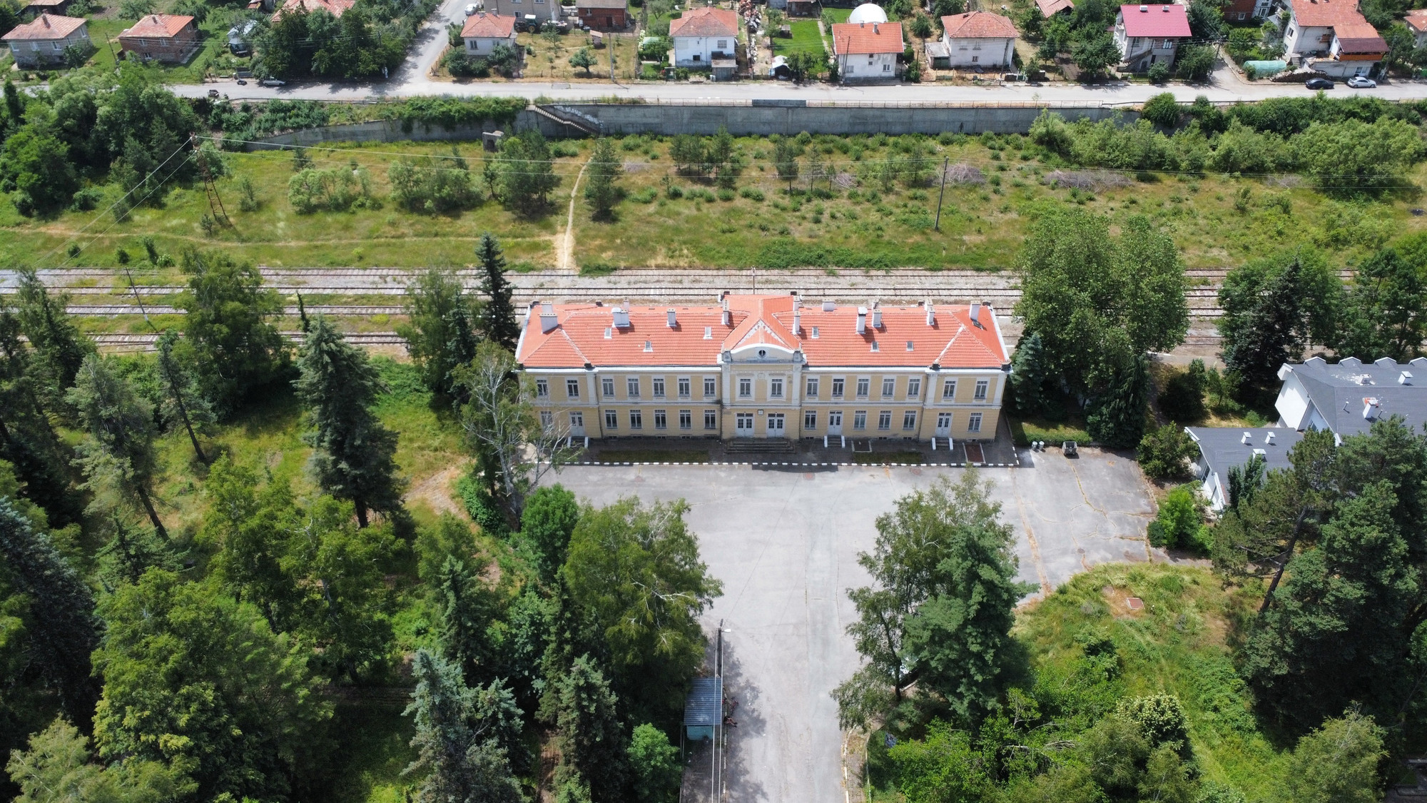 Aerial view of a large, rectangular railway station with a red-tiled roof, surrounded by greenery and trees. The building is adjacent to a railway track. Several small houses with red rooftops are visible in the background, separated from the building by the railway and open green space.
