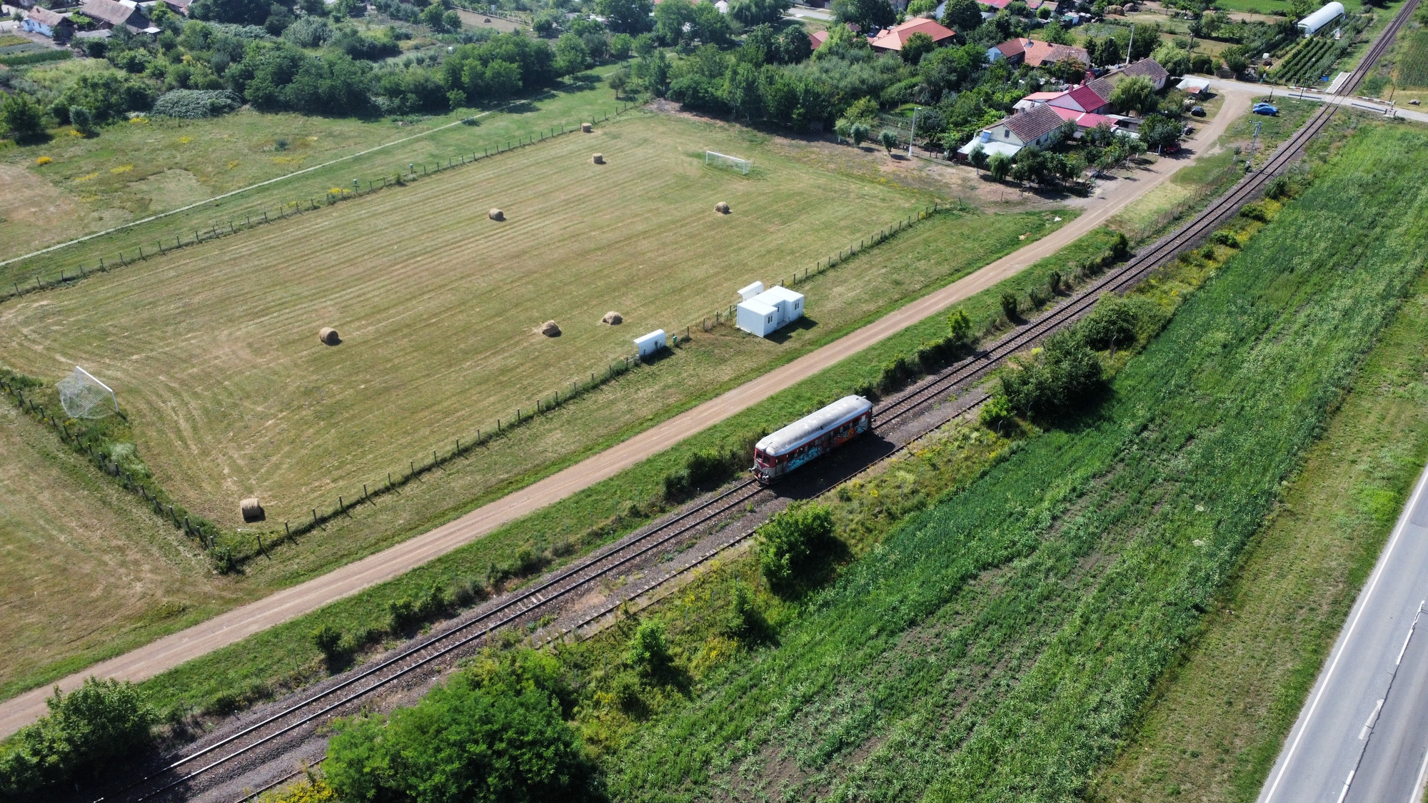 A small single carriage railbus is traveling along a railway track surrounded by lush green fields. To the left, there is a fenced soccer field with two goals and scattered hay bales. At the top of the picture there is a cluster of houses. A road runs parallel to the railway on the right side.
