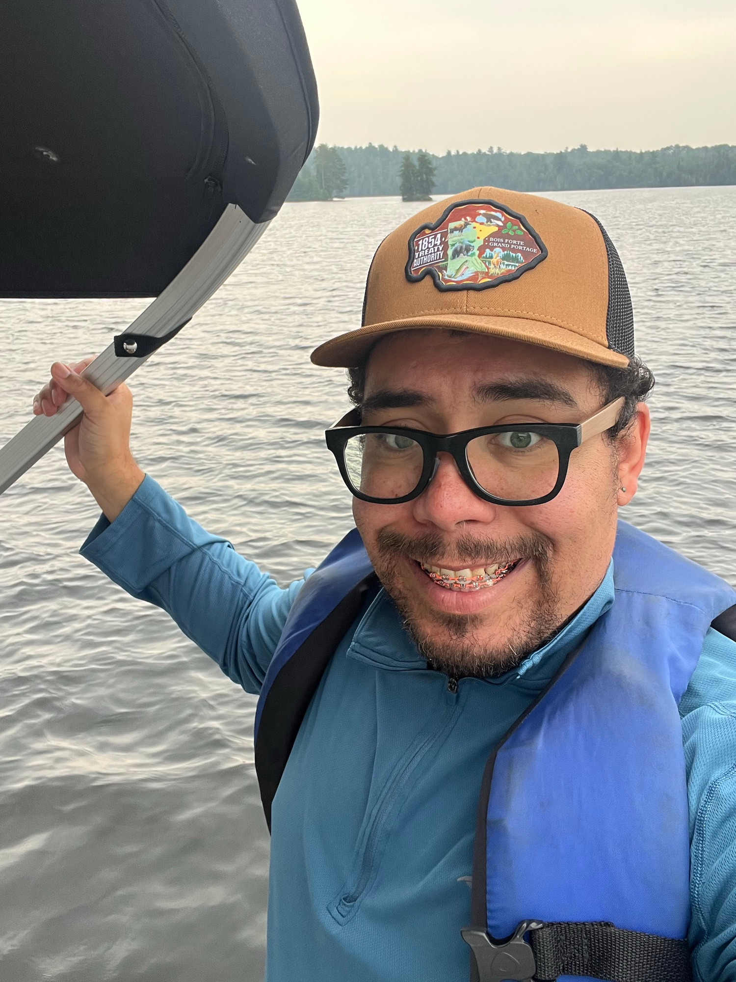 A person stands on a boat. They are wearing a tan baseball cap, a blue top, and a blue life preserver. They are looking into the camera and smiling.
