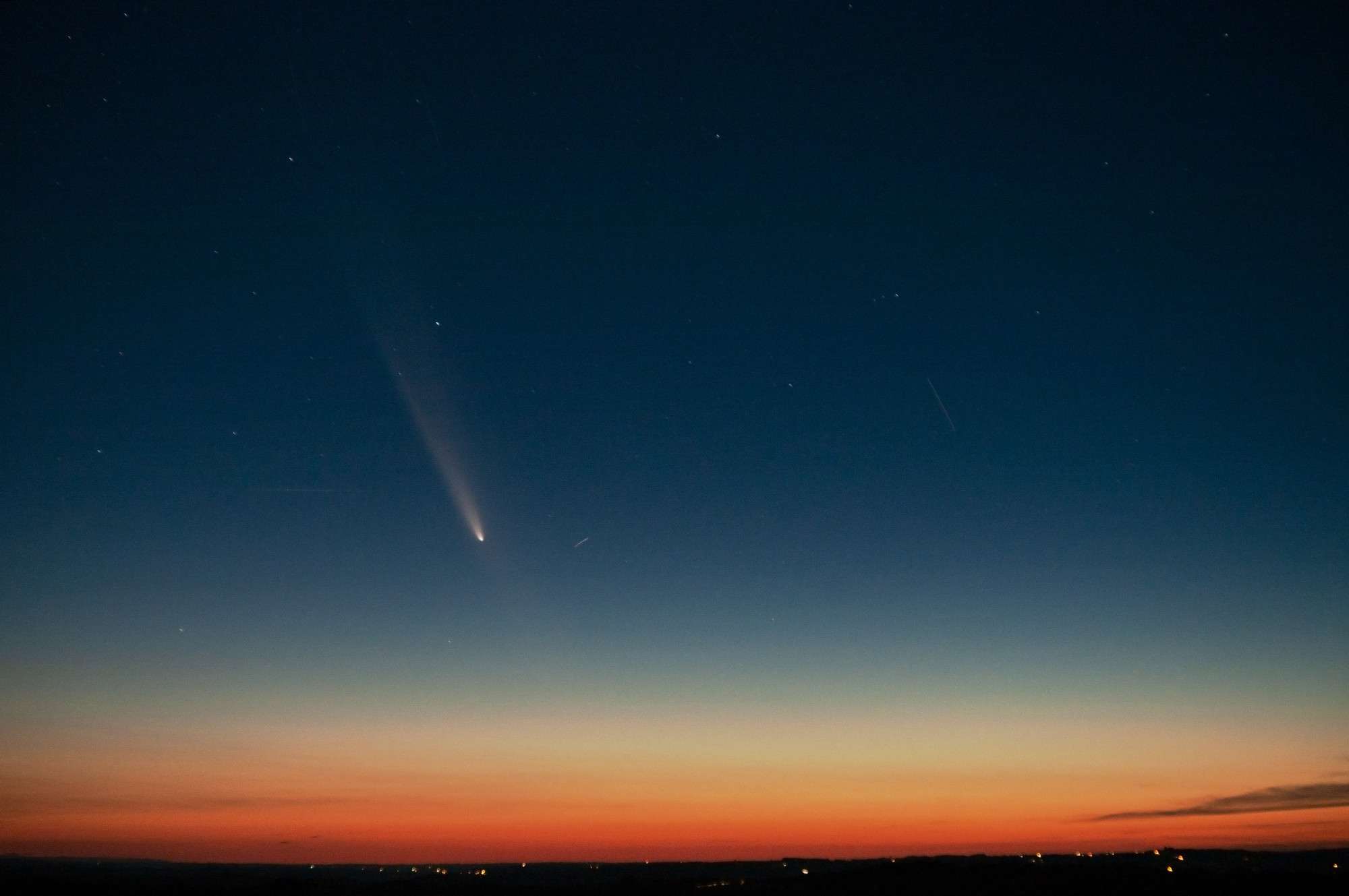 Photo prise avec quelques secondes de pose au crépuscule. On a une ligne d'horizon plate tout en bas de l'image, noire, éclairée de quelques lumières distantes. Le ciel recouvre la majeure partie de la photo, avec un dégradé, partant du bas, de rouge-orangé à bleu nuit. Quelques étoiles sont visibles. Au milieu, un peu à gauche, le ciel est traversé par la comète, qui apparaît petite dans le ciel. Avec sa traînée derrière elle elle est dirigée vers le bas.