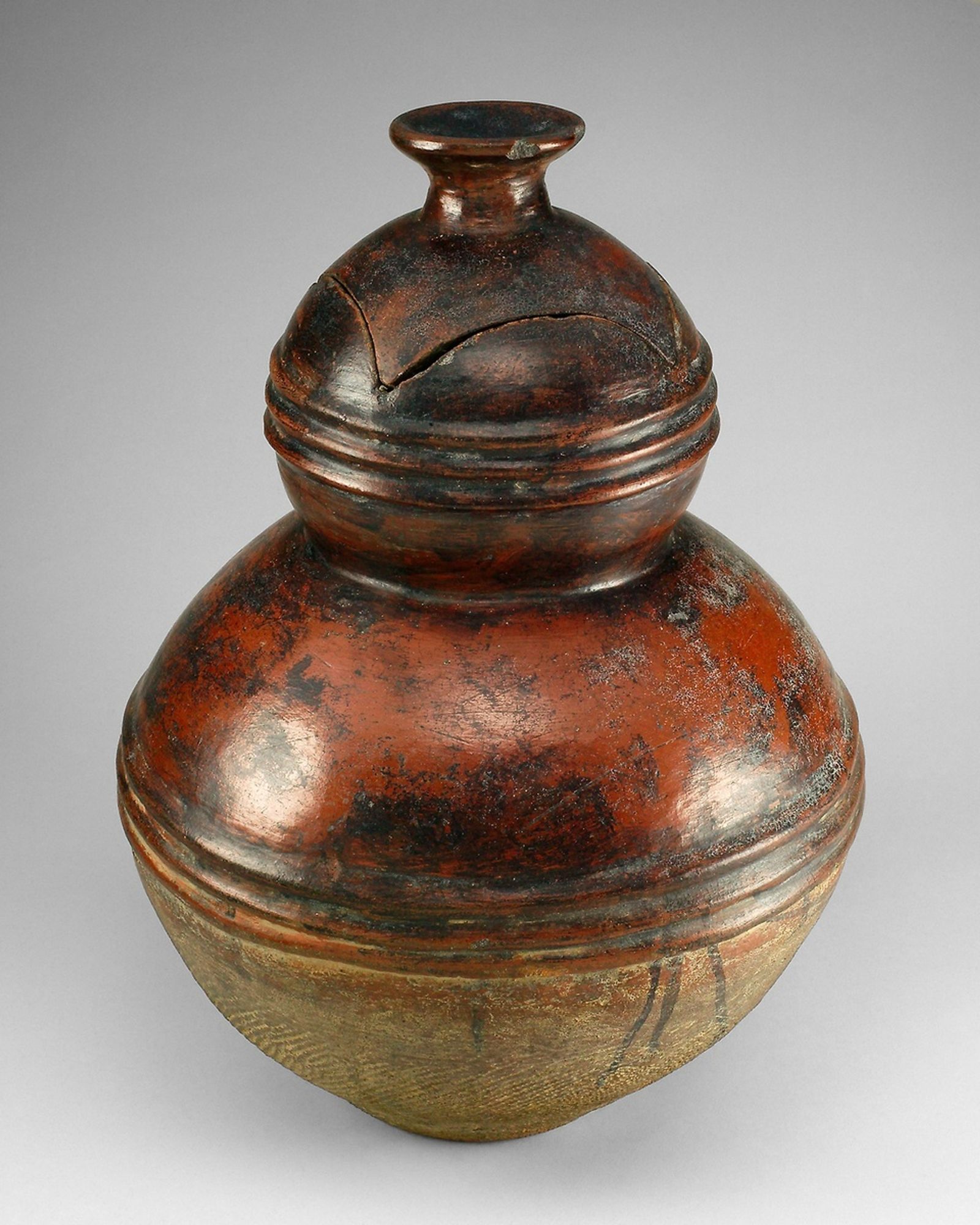 Among the Gur-speaking peoples, potters use the direct pull method, pushing into a lump of clay to form the pot’s base and pulling upward while rotating the mass to form the walls. They then scrape the clay to consolidate it and to perfect the form. Elegant, round-bodied containers such as these, which feature a lid cut seamlessly from the body and a flared topknot that acts as a handle, are made by many Gur-speaking peoples and are intended to hold valuables.
This robust vessel has two tiers that are each encircled at midpoint by a series of three ridges. A topknot handle and a cutaway lid crown the upper tier, and this, along with the upper half of the lower tier, is smoothly burnished and stained with a red slip that has partially blackened. In contrast, the bottom half of the lower tier was consolidated with a roulette or corncob, giving it a textured surface. According to Douglas Dawson, the Nuna specialize in these two-tiered forms.
While the valuables stored within such containers may include money and jewelry, their worth may also be defined more esoterically. For example, clans, lineages, and individuals possess objects that are treasured for their role in forging a connection with the vital forces of nature.  According to scholar Christopher Roy, these may include “animal skulls and tails, rings, amulets, bracelets, stools, bottles, and anthropomorphic figures in clay and wood.”  [See also 2005.232].

Gift of Keith Achepohl