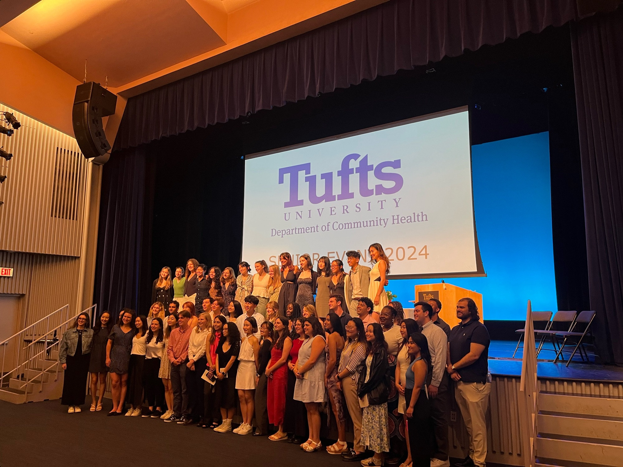 A group of senior undergraduates are posed for a group photo on an auditorium stage. A banner for Tufts University’s Department of Community Health appears behind them.