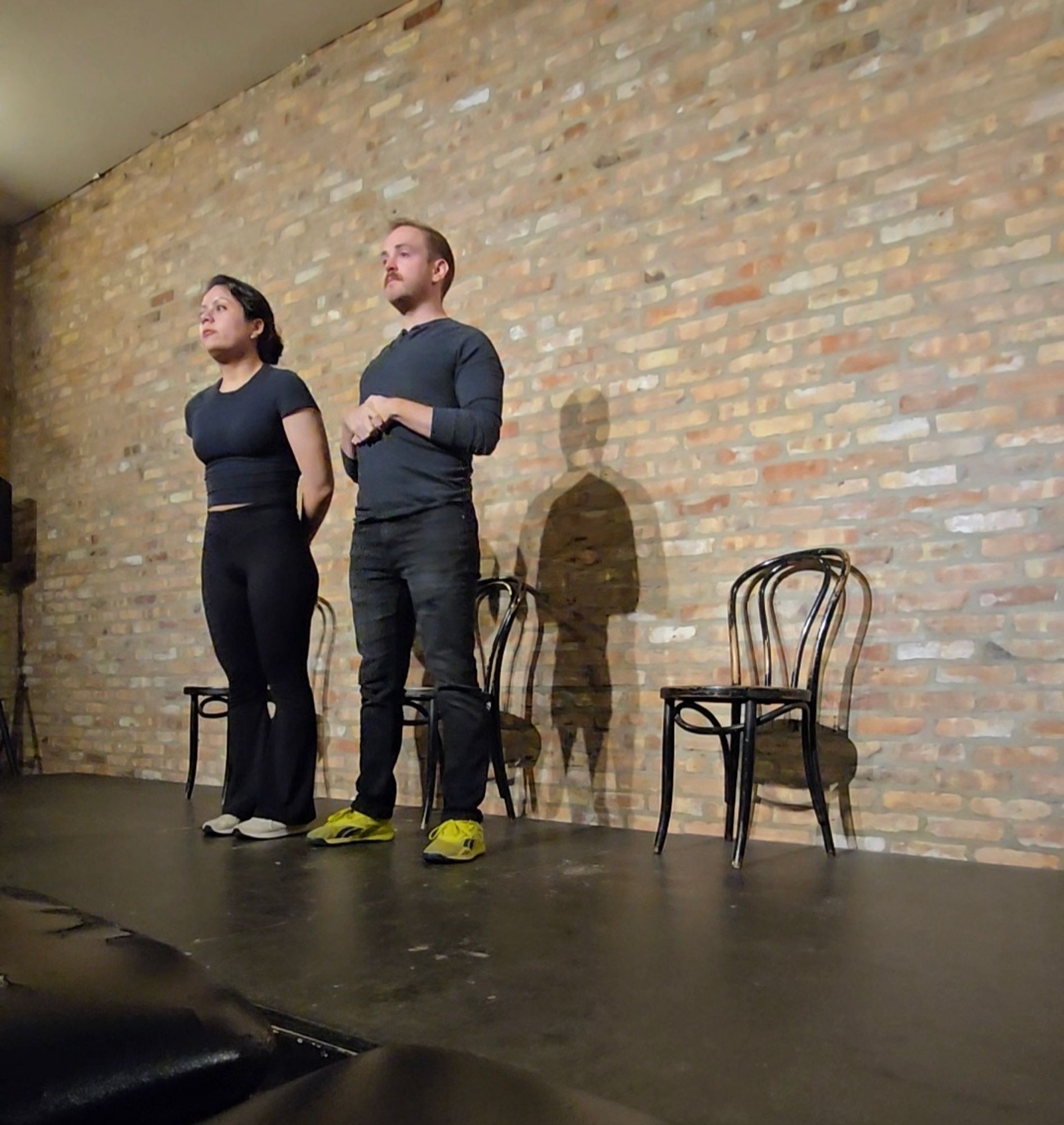 Two people dressed in black, on stage. They stand in front of a brick wall.