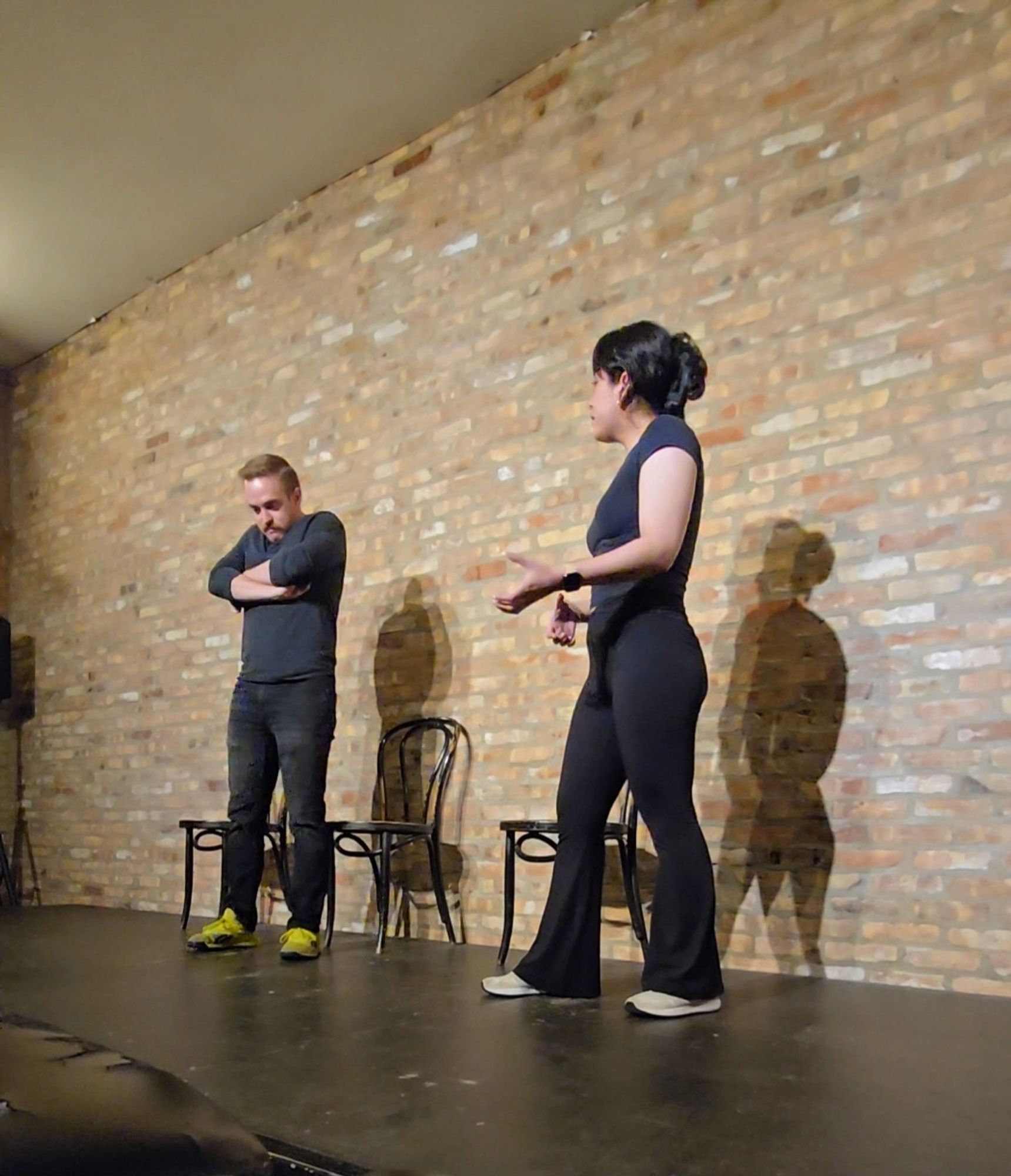 Two people dressed in black, on stage. They stand in front of a brick wall. Jake is on the left with his arms crossed, looking grumpy.