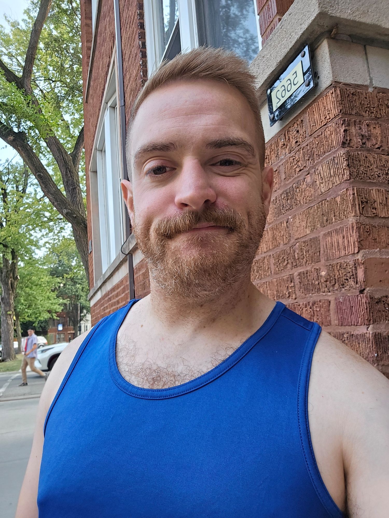 Selfie of Jake with a haircut, wearing a blue tank top. He is outside of a brick house