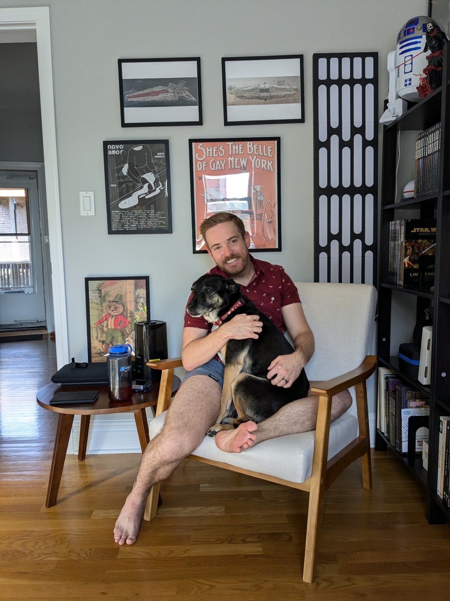 Jake and his dog freyja sitting together in a chair. He is in shorts and a red shirt. The dog is looking away from the camera