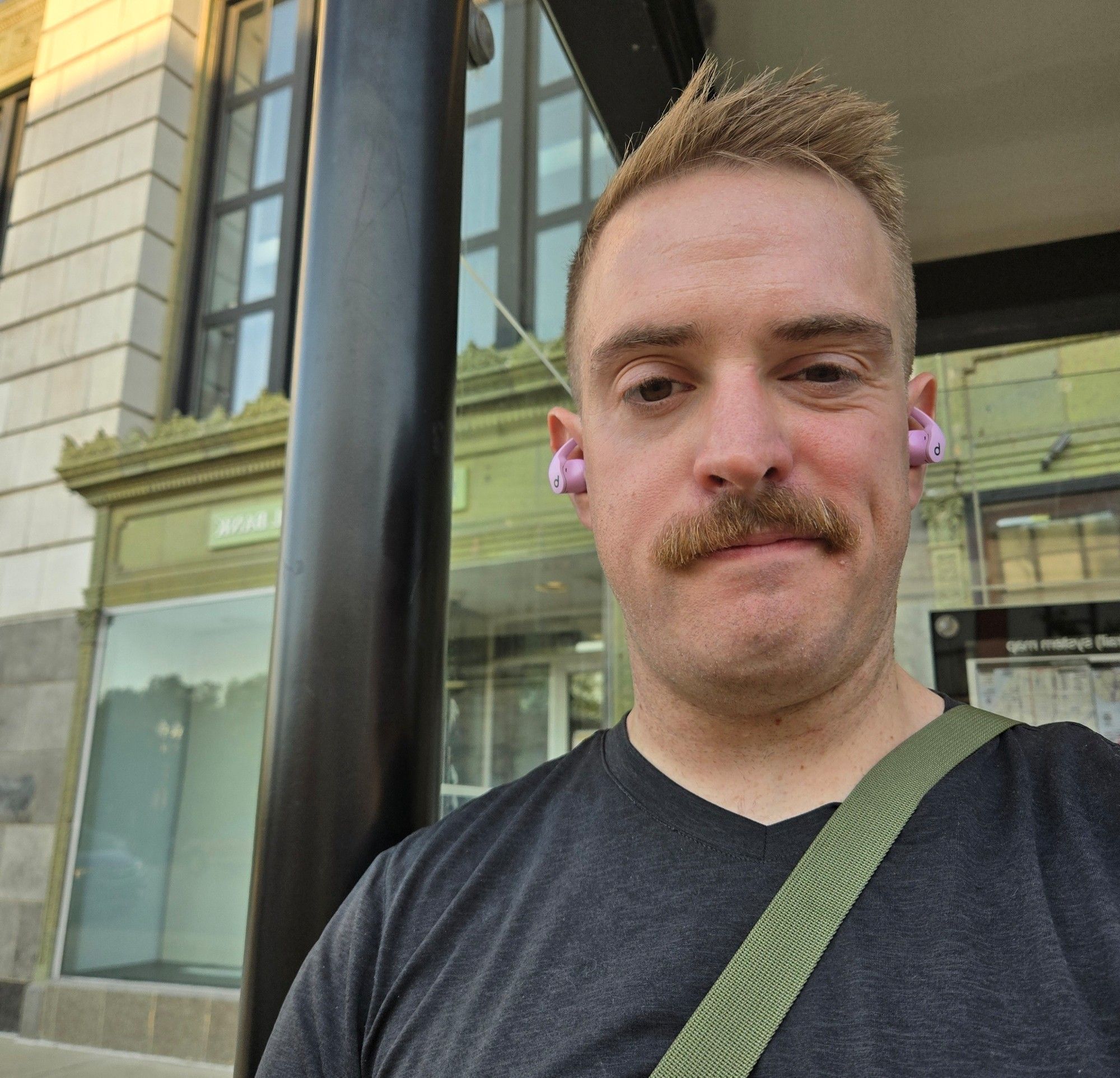 Selfie of Jake with a new haircut and mustache. He is at a bus station.