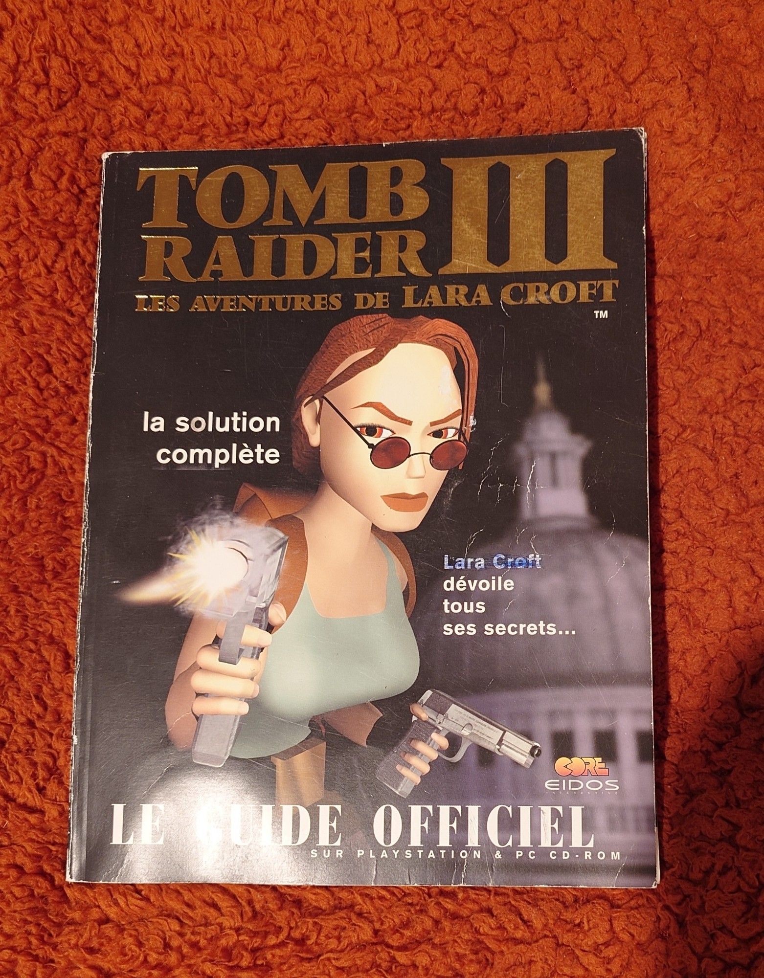 Couverture du guide officiel Tomb Raider 3 sortie en 1998. Les coins sont un peu abîmés, la couverture est un peu cornée. Quelqu'un à colorer le nom Lara Croft avec un stylo bleu.