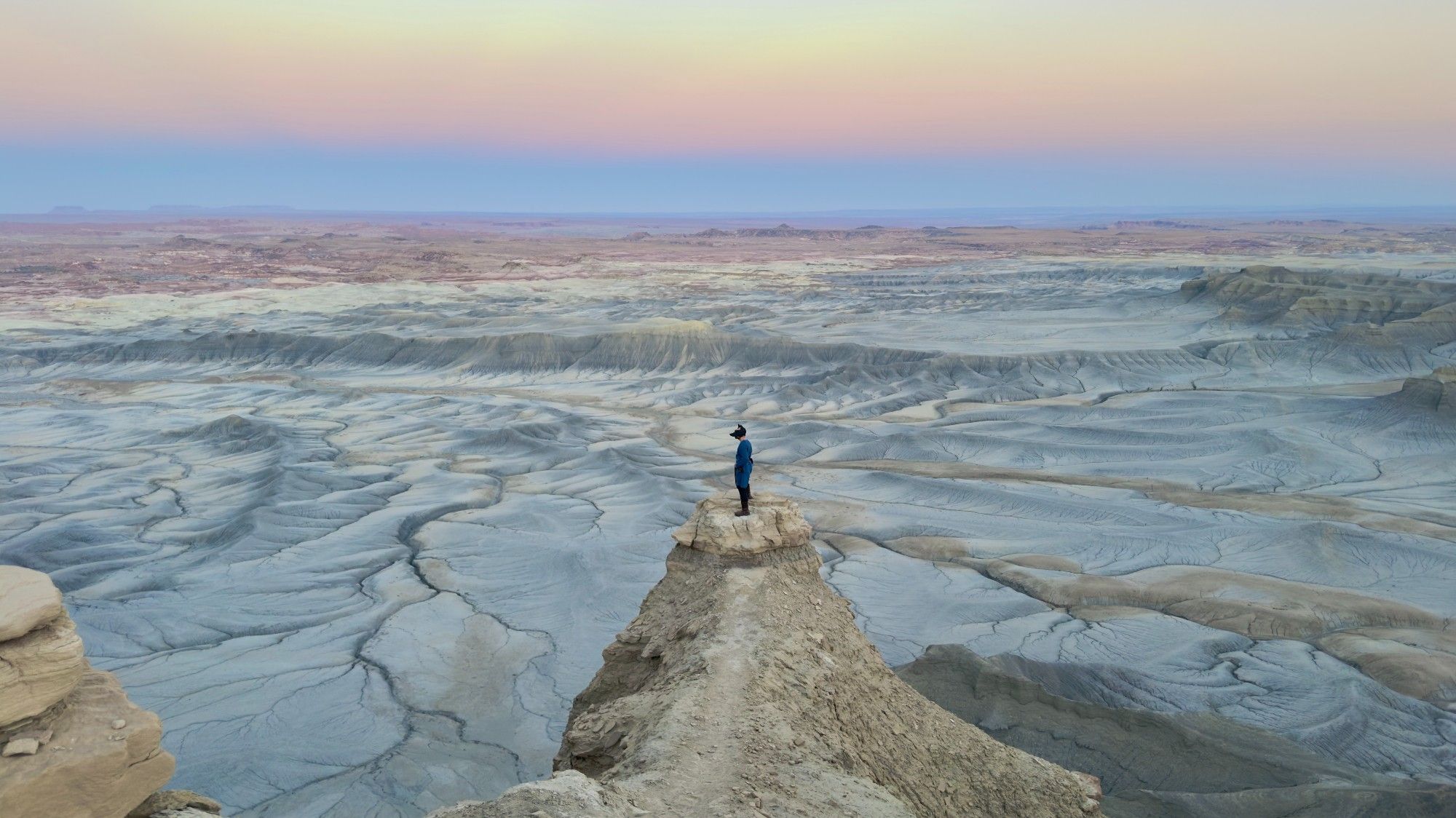 Cosplaying as Lago Vaari from the Noss Saga while standing on a precarious point of sandstone a thousand feet over a gray landscape of eroded sediments, with pink and lavender colors stretching in the sky.