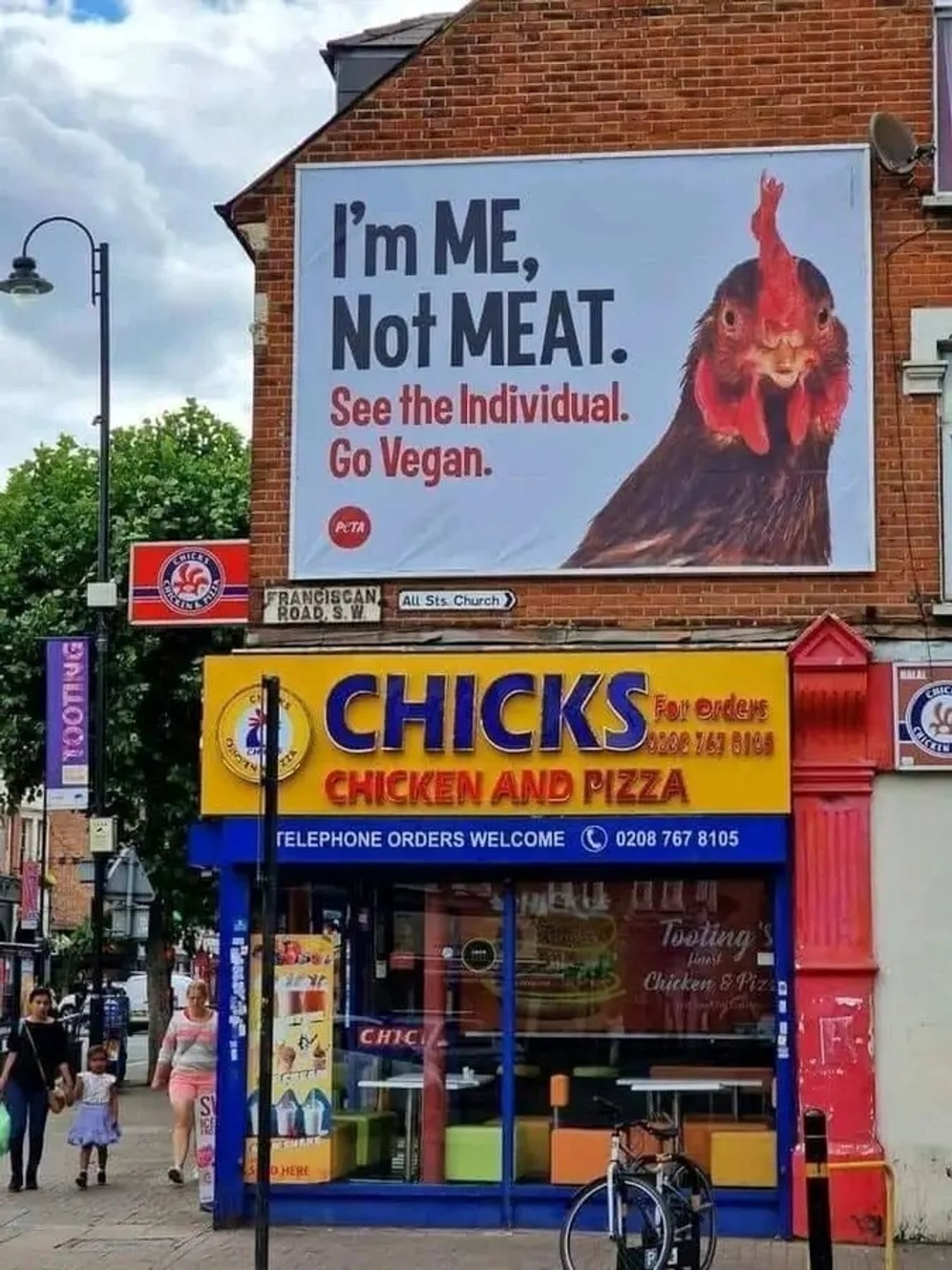 Foto eines Hühnerfleisch-Fast-Food-Imbisses, über dem ein großes Plakat an der Hauswand angebracht ist.
Darauf ist ein Huhn abgebildet samt Text: 
"I'm me, not meat.
See the individual.
Go vegan."