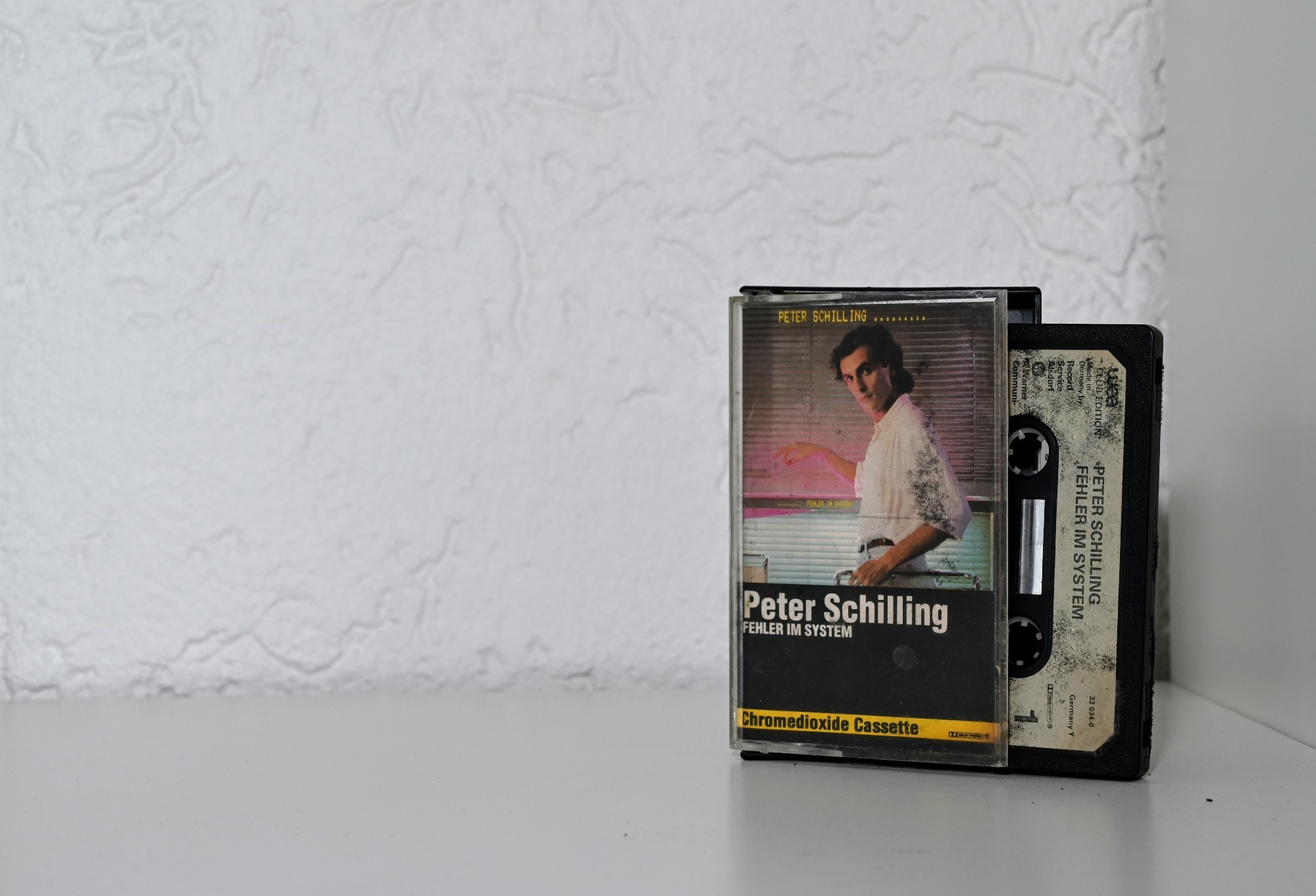 Musik-Kassette vor weißem Hintergrund stehend. Die Hülle ist halb geöffnet, die Kassette etwa halb herausgezogen.
Das Cover zeigt im Halbprofil eine schwarzhaarige dünne Person in einem weißen Hemd vor einer Jalousie. Unter der Abbildung eine weiße Schrift auf schwarzem Hintergrund: "Peter Schilling",  "Fehler im System".