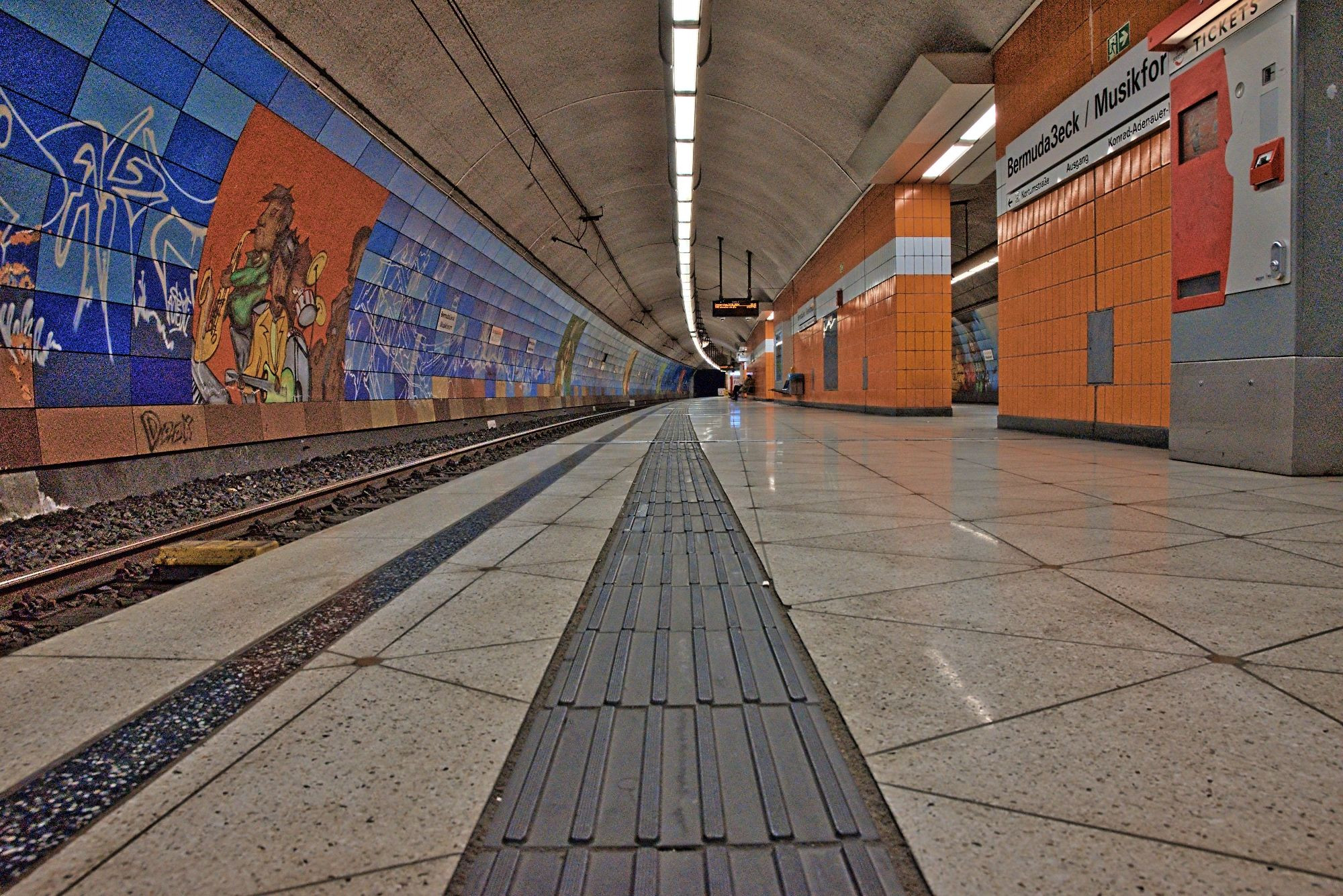 Menschenleerer U-Bahnhof, Froschperspektive, den Bahnsteig entlang. Zentral ein dunkler Blindenleitstreifen auf hellen Fliesen. Links der Gleiskörper, gekachelte Seitenwand mit künstlerischen Graffiti. Rechts ein Fahrscheinautomat und orange gekachelte Mittelwände. Die Decke ist gewölbt und besteht aus Sichtbeton