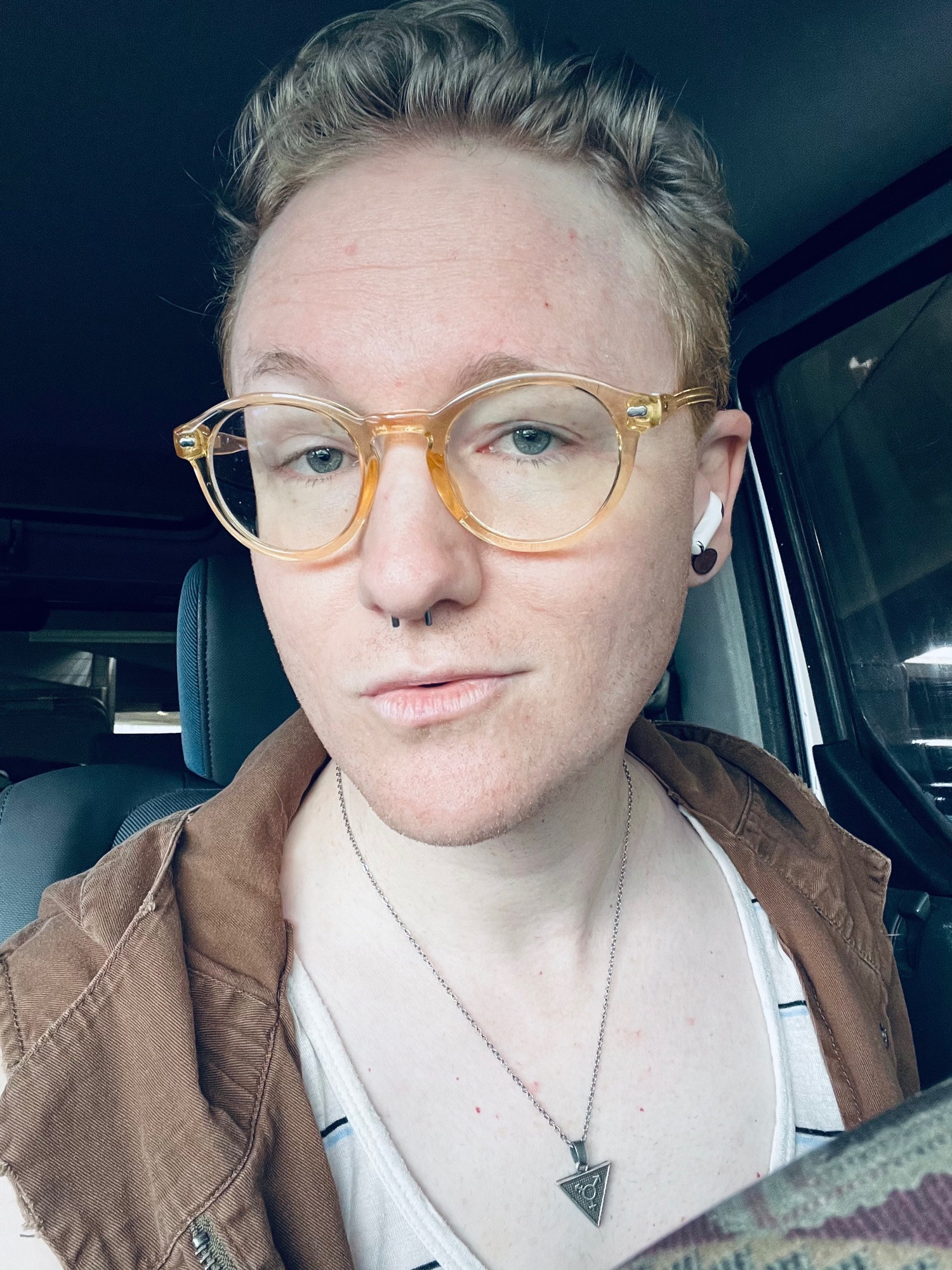 Fox in its car wearing a white scoop neck top with horizontal black and white stripes, a sleeveless brown hooded cotton jacket, a small silver chain with a triangular pewter pendant with the transgender symbol on it, and clear yellow glasses. her curly gray hair is pulled back in a low pompadour. fæ is looking into the camera with a fleeting half-smile.