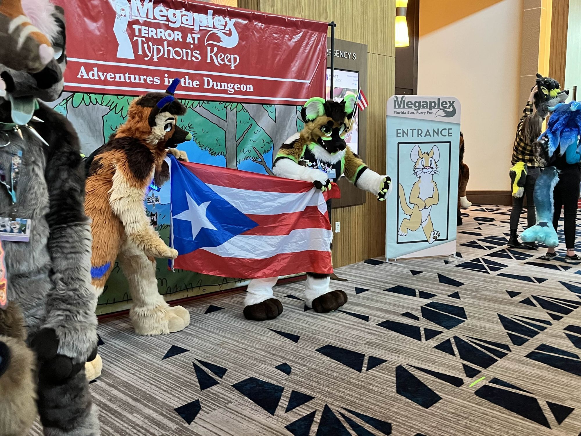 Two fursuiters in frame, both are holding the Puerto Rican flag in front of a themed banner for the Megaplex 2024 furry convention. Both fursuiters are brown with colored accents, the one on the left has blue accents while the one on the right is lime green.