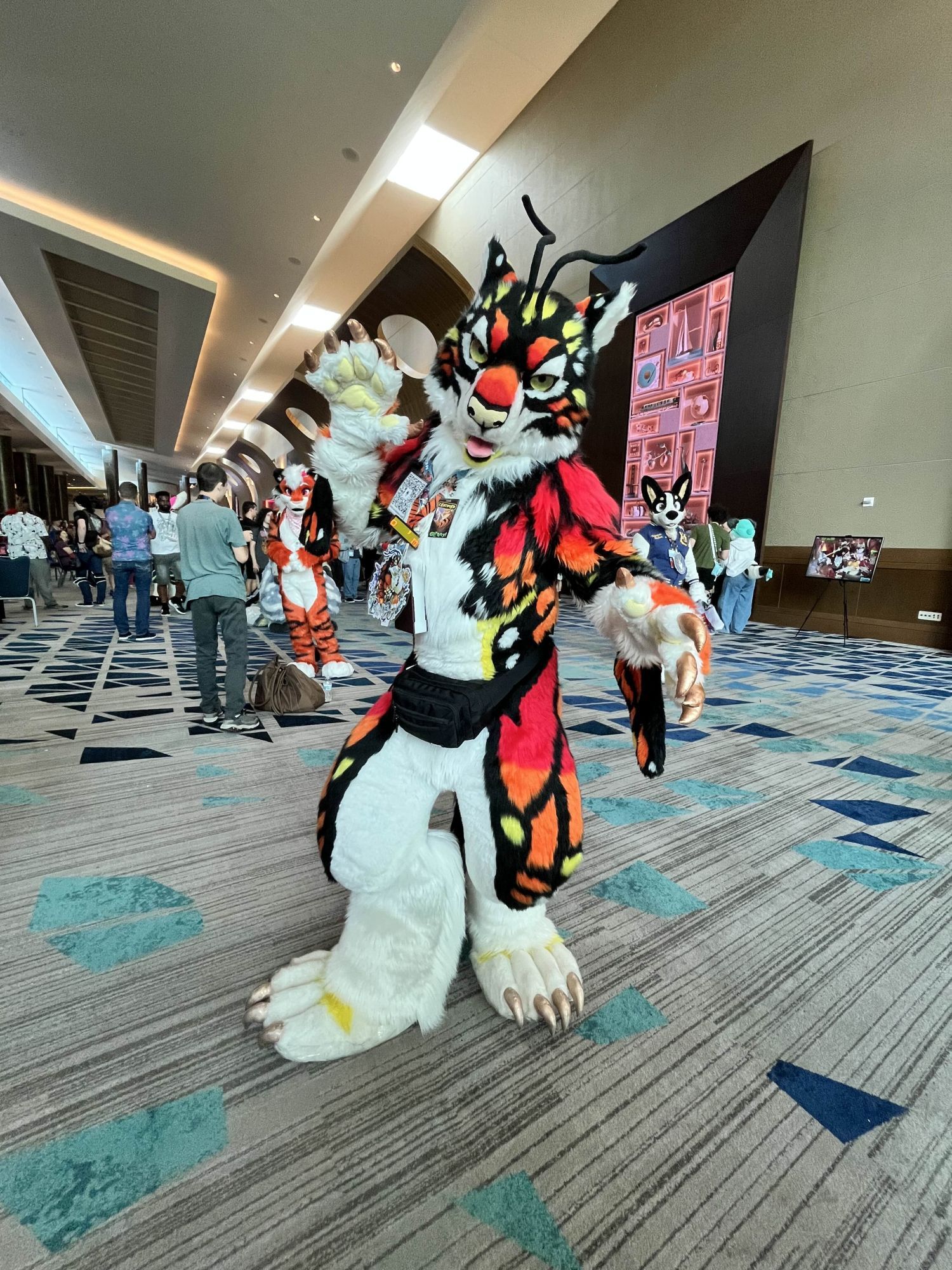 A photo of a fursuiter at the Megaplex 2024 furry convention. The suit has black, white, red, orange, and yellow colors in the pattern that evokes the appearance of a monarch butterfly with a feline appearance for the head.