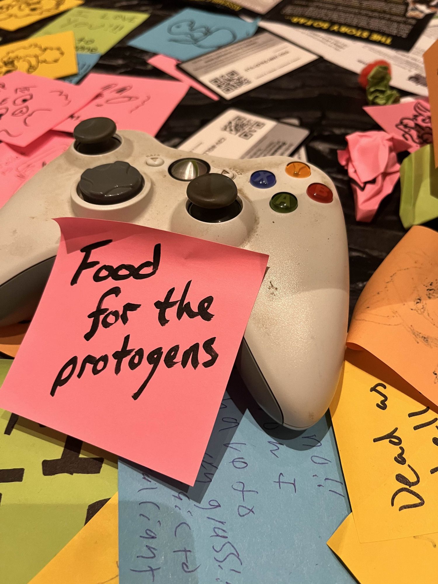 A well worn Xbox 360 controller on a table surrounded by sticker notes expressing sorrow over its demise. The main sticky note shown reads "Food for the protogens"