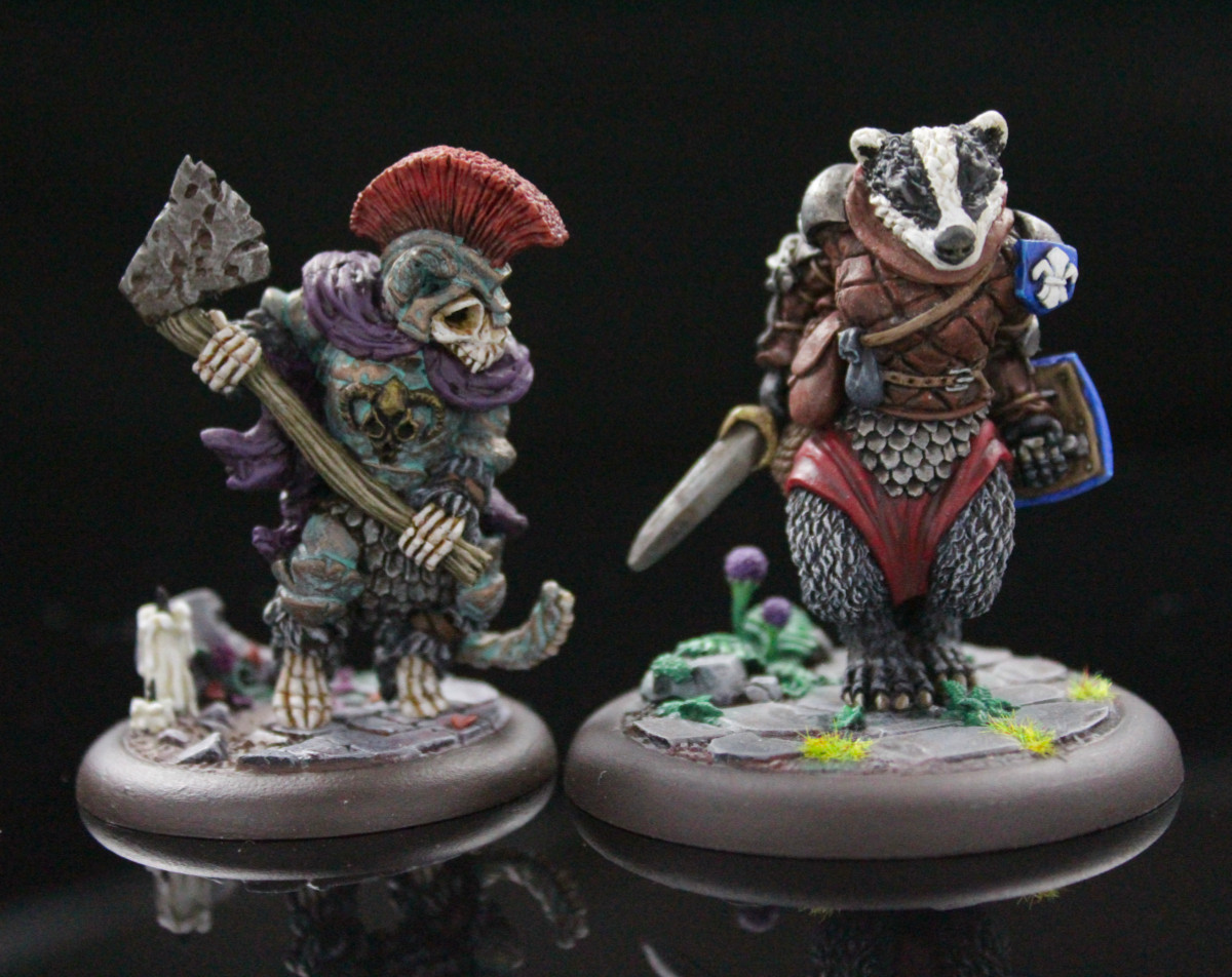 A pair of minis from the game Burrows & Badgers. Left is an undead fox warrior with an axe, he is nothing but bones in armor and very grody. Right is a badger warrioress with sword and shield, she is much cleaner except for a normal amount of badger grime.