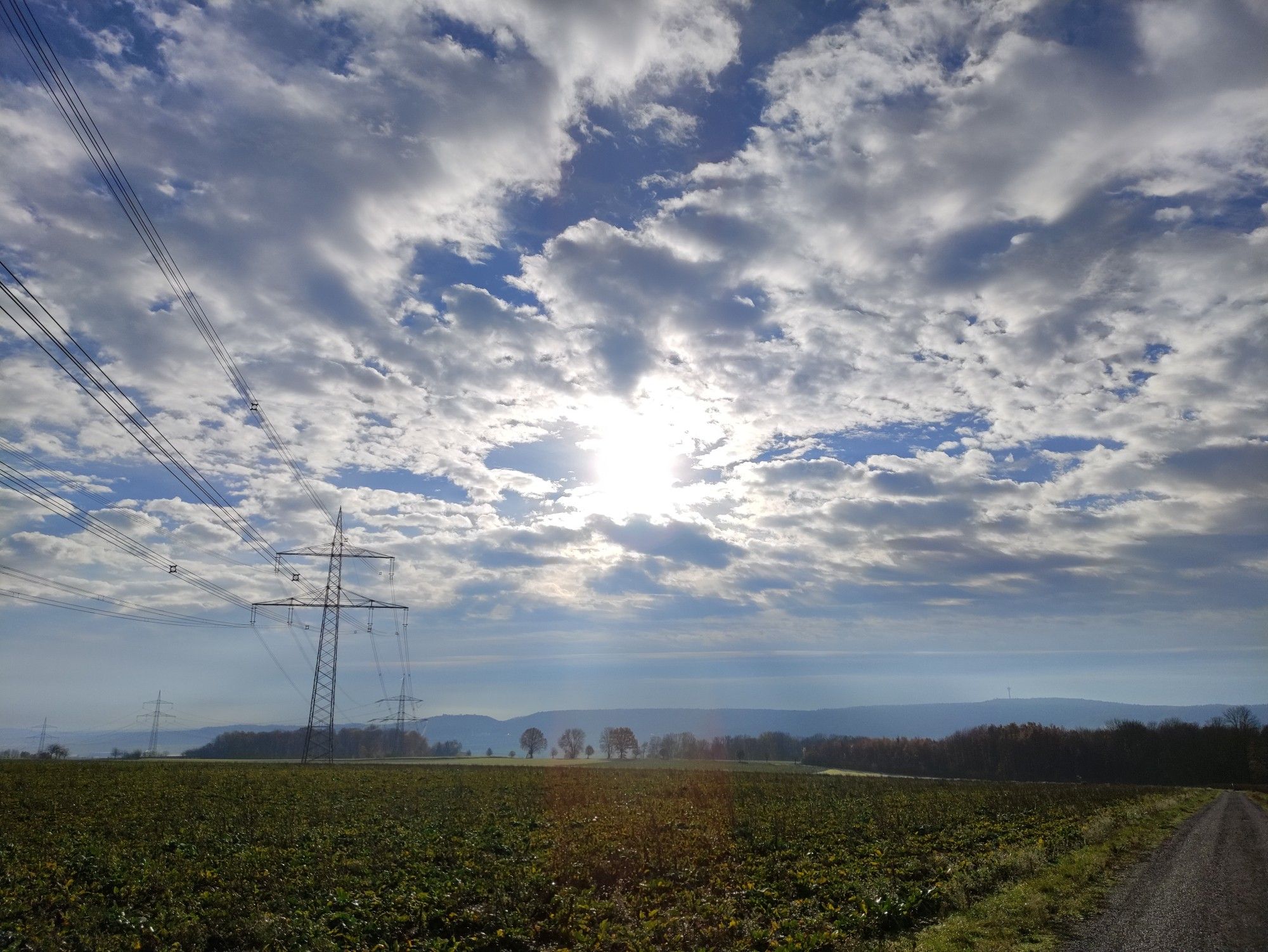 Felder, Bäume, Wald, Strommasten, darüber größtenteils bedeckter Himmel mit blauen Durchbrüchen und Sonne