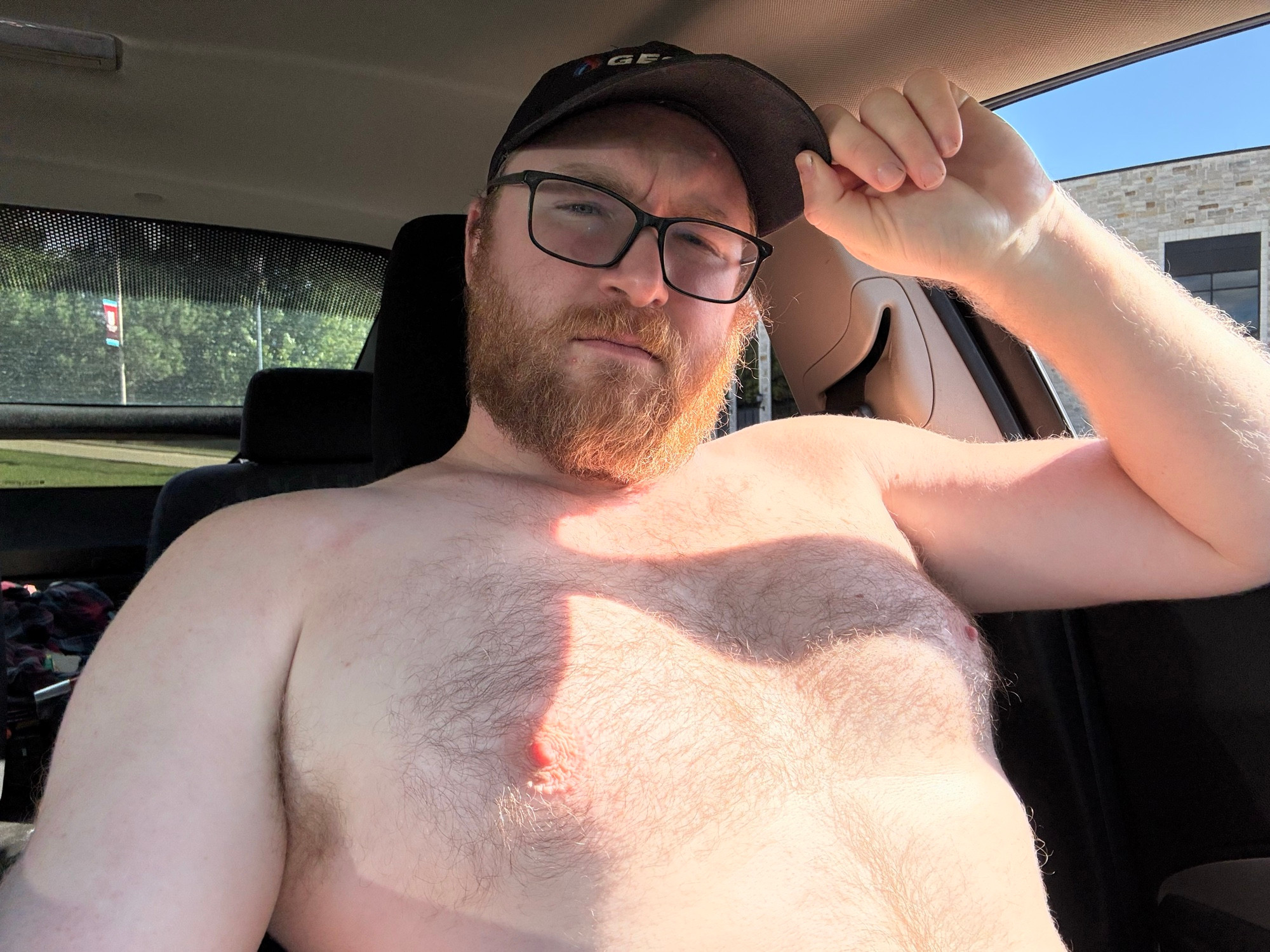 Bearded, bespectacled, bare chested man in the drivers seat of a car. 