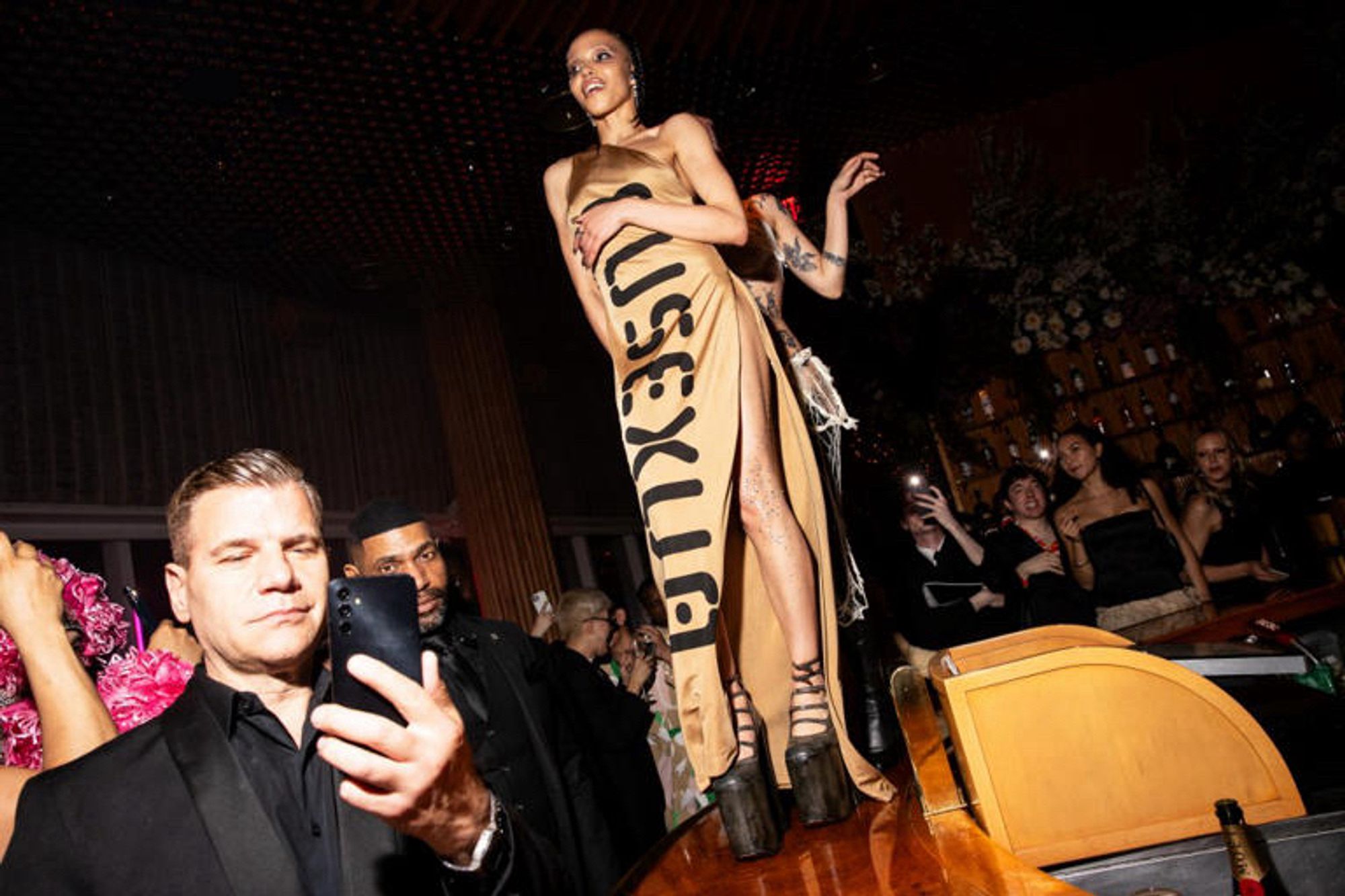 FKA Twigs standing on a bar during her performance at a MET Gala afterparty. Wearing a camel colored one-shoulder tunic dress with the text “EUSEXUA” (title of her forthcoming album). She also dons a earthy, dark brown pair of Marc Jacobs Kiki boots