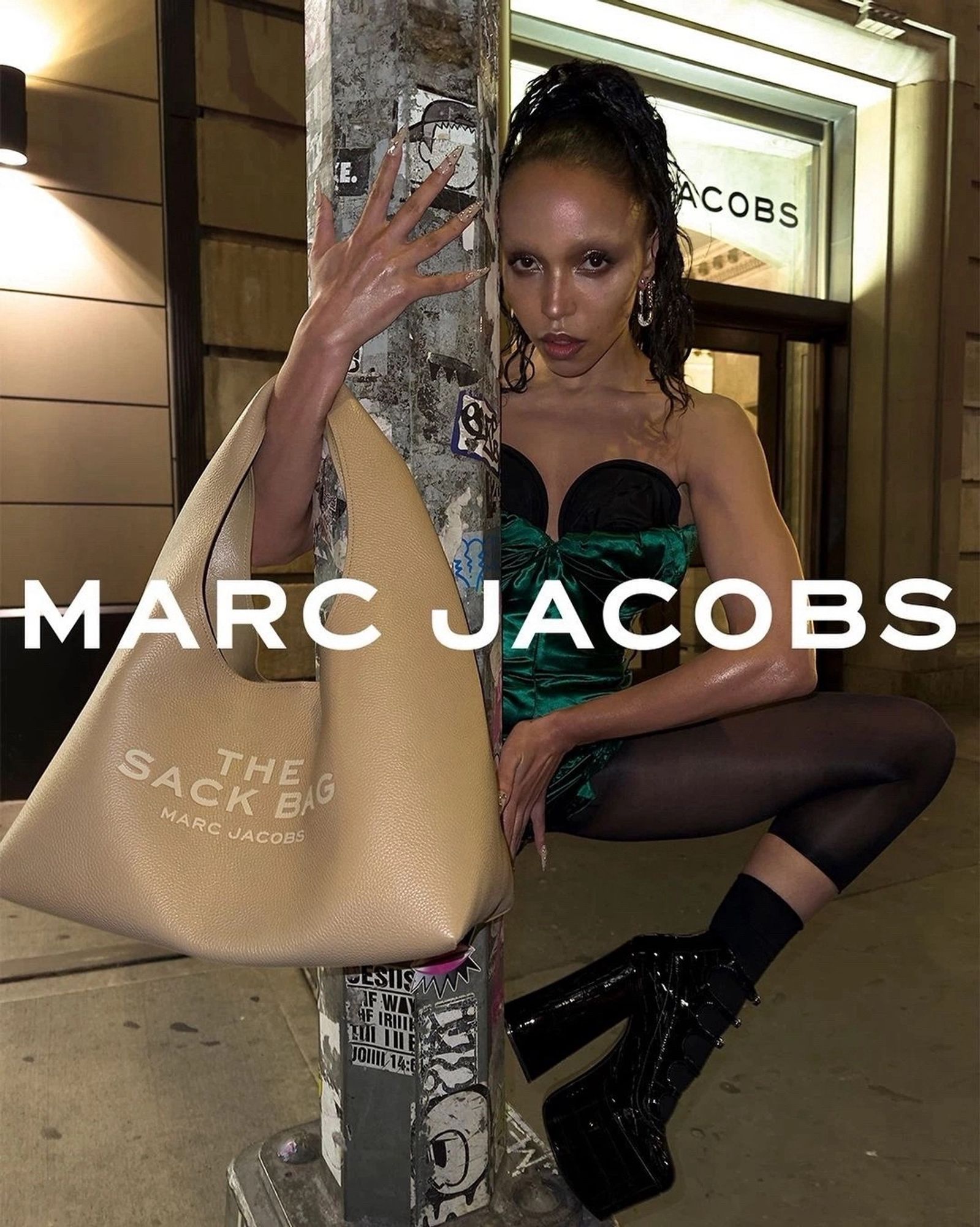 FKA Twigs posing with an arm around a lightpost, showing off the Marc Jacobs “Sack” bag and black Kiki ankle boots outside the Marc Jacobs headquarters in Soho