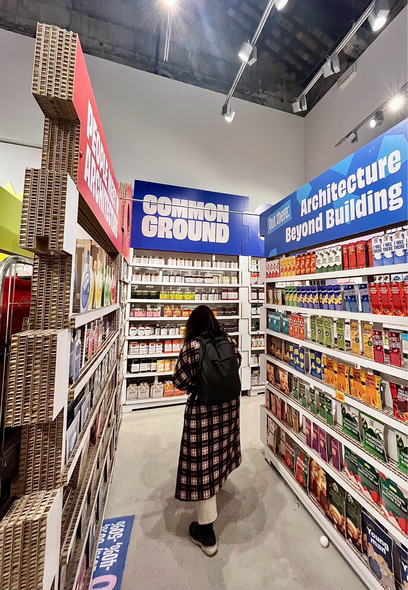 ein supermarkt aus pappkartonregalen & bildern statt ware. eine junge frau steht in der mitte zwischen den regalen. von oben über lichtleisten beleuchtet.