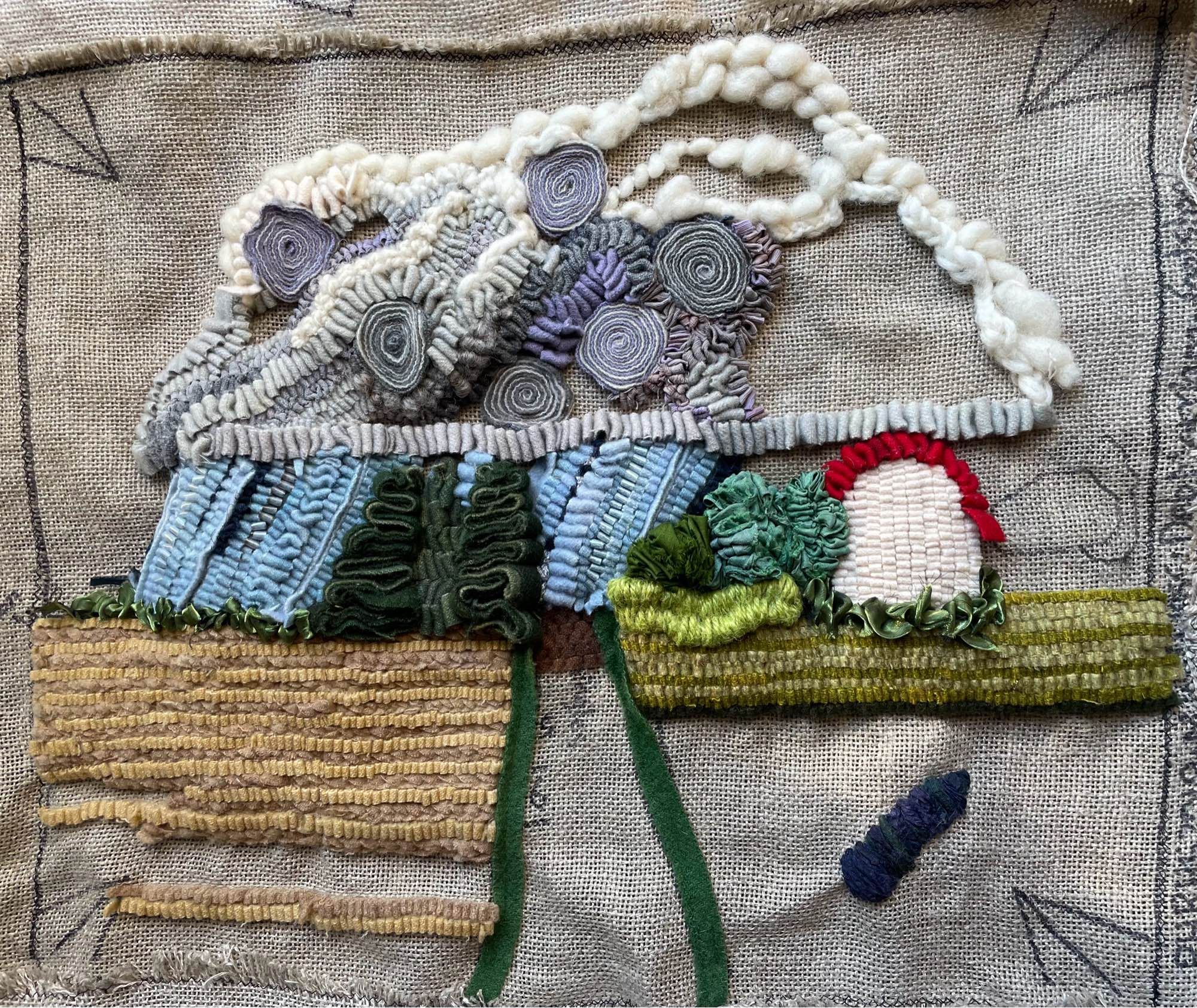 Rug hooked work in progress of hand-dyed wool flannel fabric strips, sari silk ribbon, twine and wool roving on linen backing, depicting a flat rural Prairie road with crop fields on each side (beige and yellow-green with a teal slough), a treeline (darker blue- greens) with a white barn on the horizon, and the distant sky filled with a huge grey-and-violet cumulonimbus cloud that’s dumping rain. Techniques used include the standard rug hooking loop stitch, reverse brick stitch, tambour stitch, couching, satin stitch, rip-and-wrinkle, quilling, stitched standing wool,and clear elastic overlay.