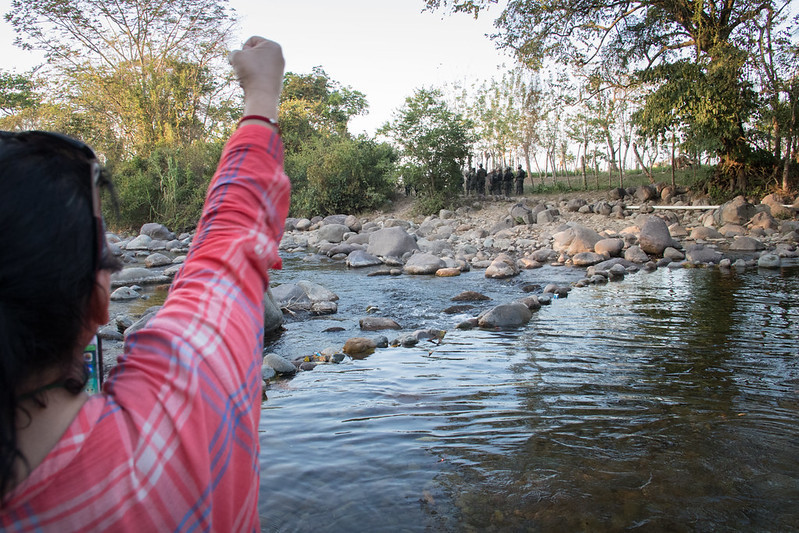 Activisten bij de Guapinol-rivier. Aan de overkant een legereenheid die bekend staat vanwege mensenrechtenschendingen. Achter hen het hek van een mijnbouwgebied. (Foto: Peg Hunter op Flickr/CC BY-NC-ND 2.0 Deed niets veranderd.)