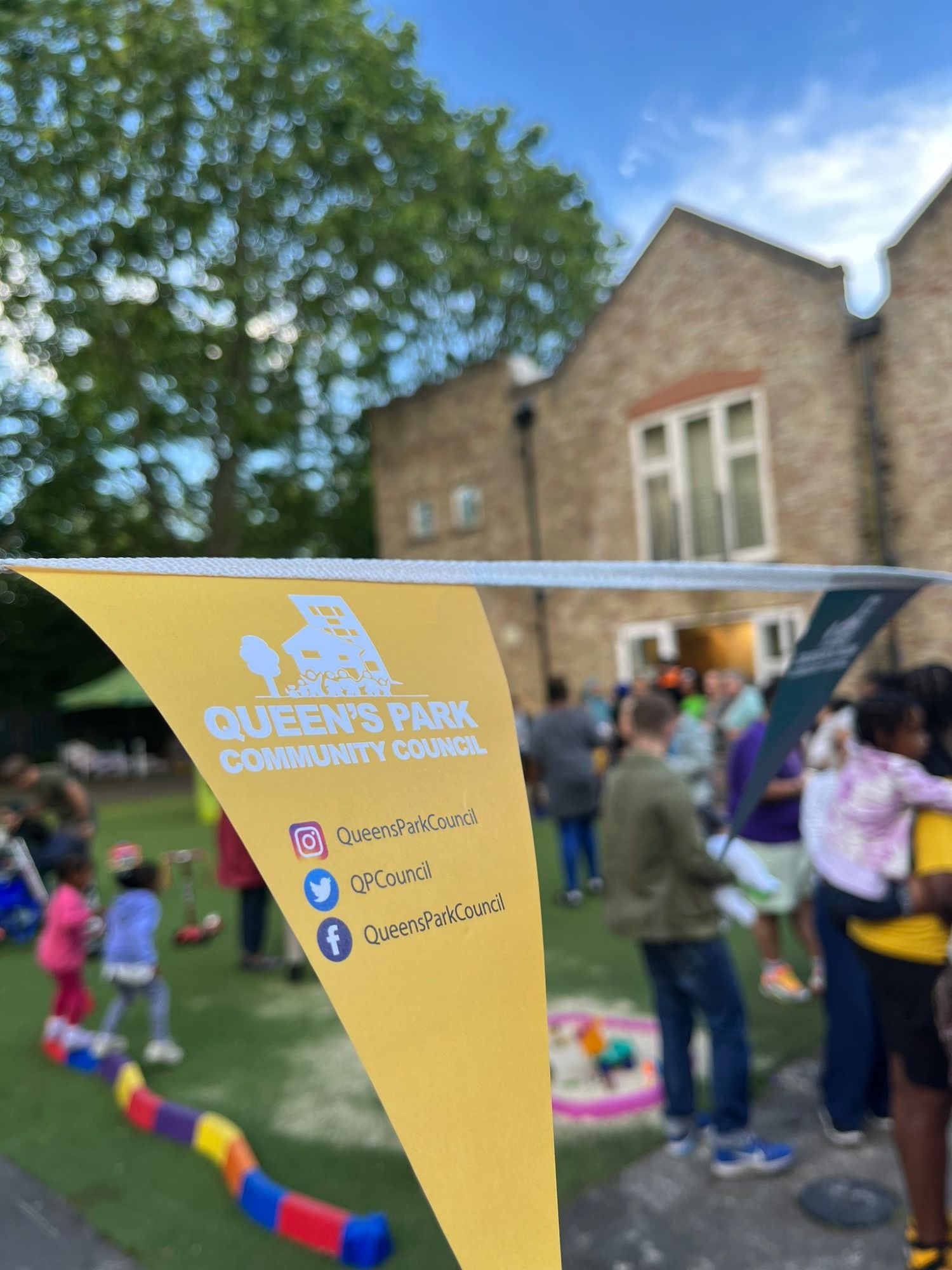 A photo with buildings and people in the background, and a bunting flag in the foreground. On the yellow bunting is the Queen’s Park Community Council logo and social media handles.