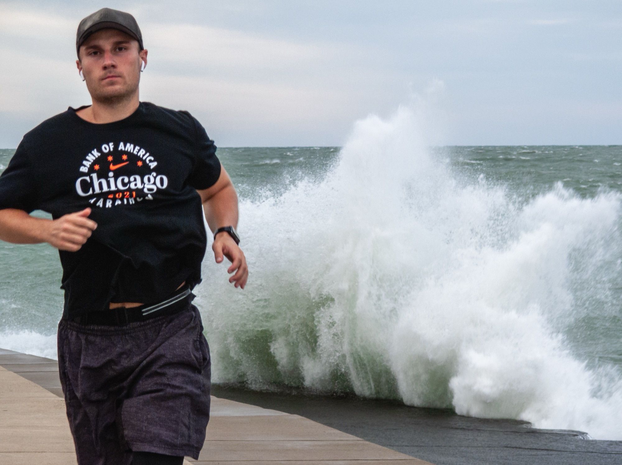 Daytime. A runner (frame right) jogs along the concrete landing off Fullteron, north of the North Avenue Beach, while a large wave from Lake Michigan splashes dramatically in the background. The runner is looking directly into the camera. The sky is slightly overcast. Colors are stylized.