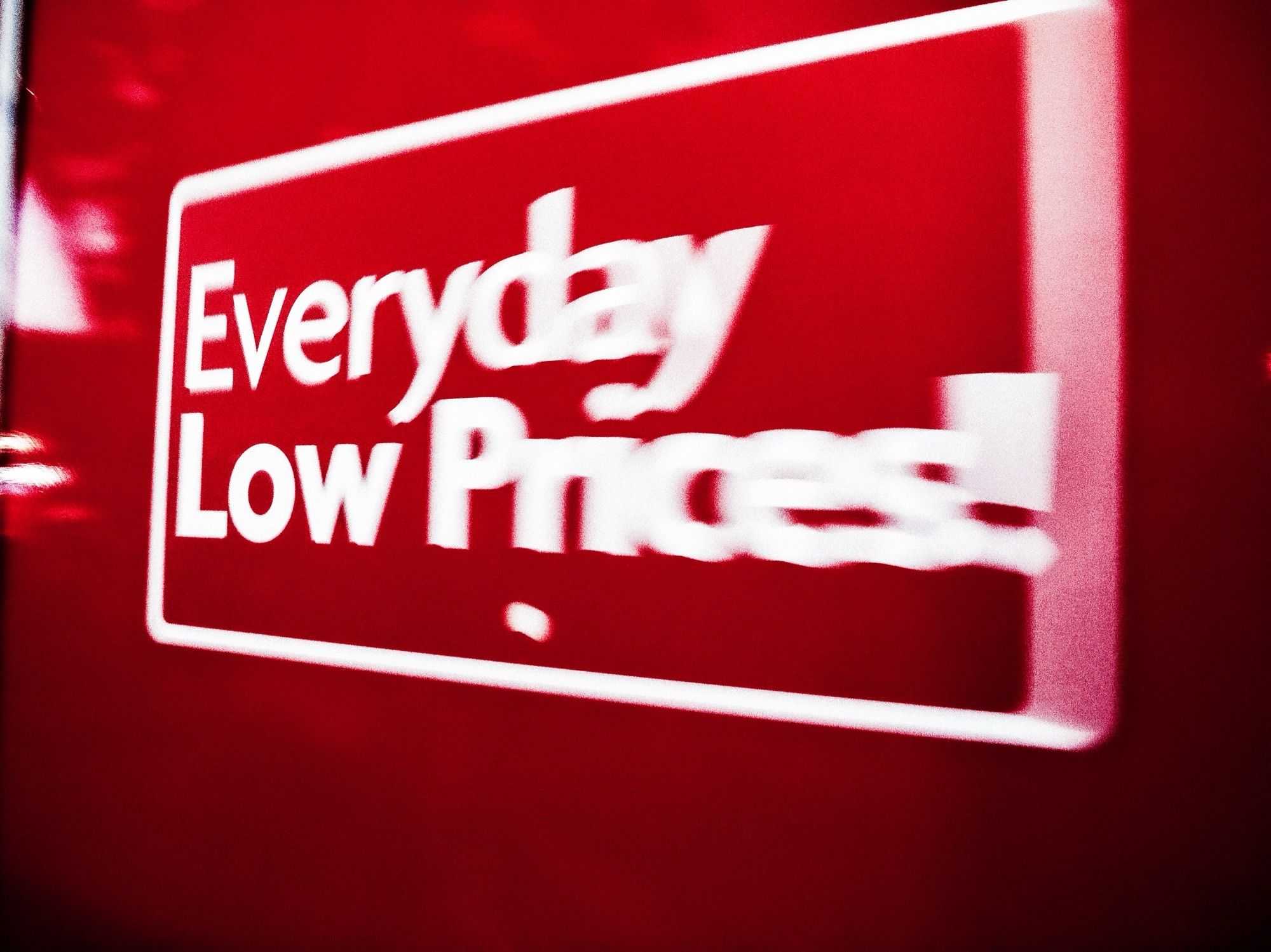 Nighttime. A sign on a Family Dollar indicating “Everyday Low Prices!”. The background is red, and the text is white. Some slight blurring caused by a slower shutter speed, and slight camera movement. Colors and luminance contrast are high, and stylized.