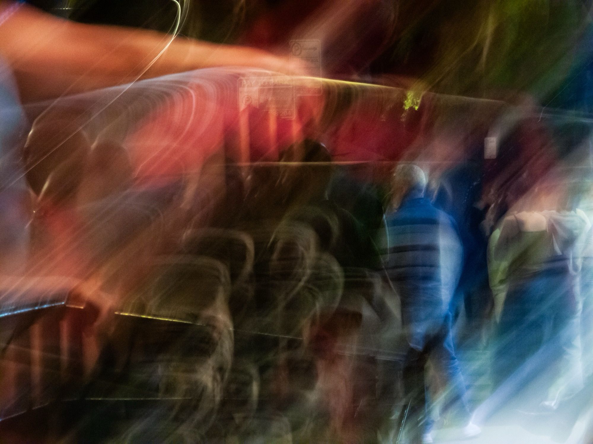 Nighttime. A man in a blue vest stands out in an abstracted street scene. Other people can be vaguely recognized standing near him. Extreme streaking and light trails are caused by a long shutter speed and intentional camera movement. Luminance contrast is high and colors are highly stylized.