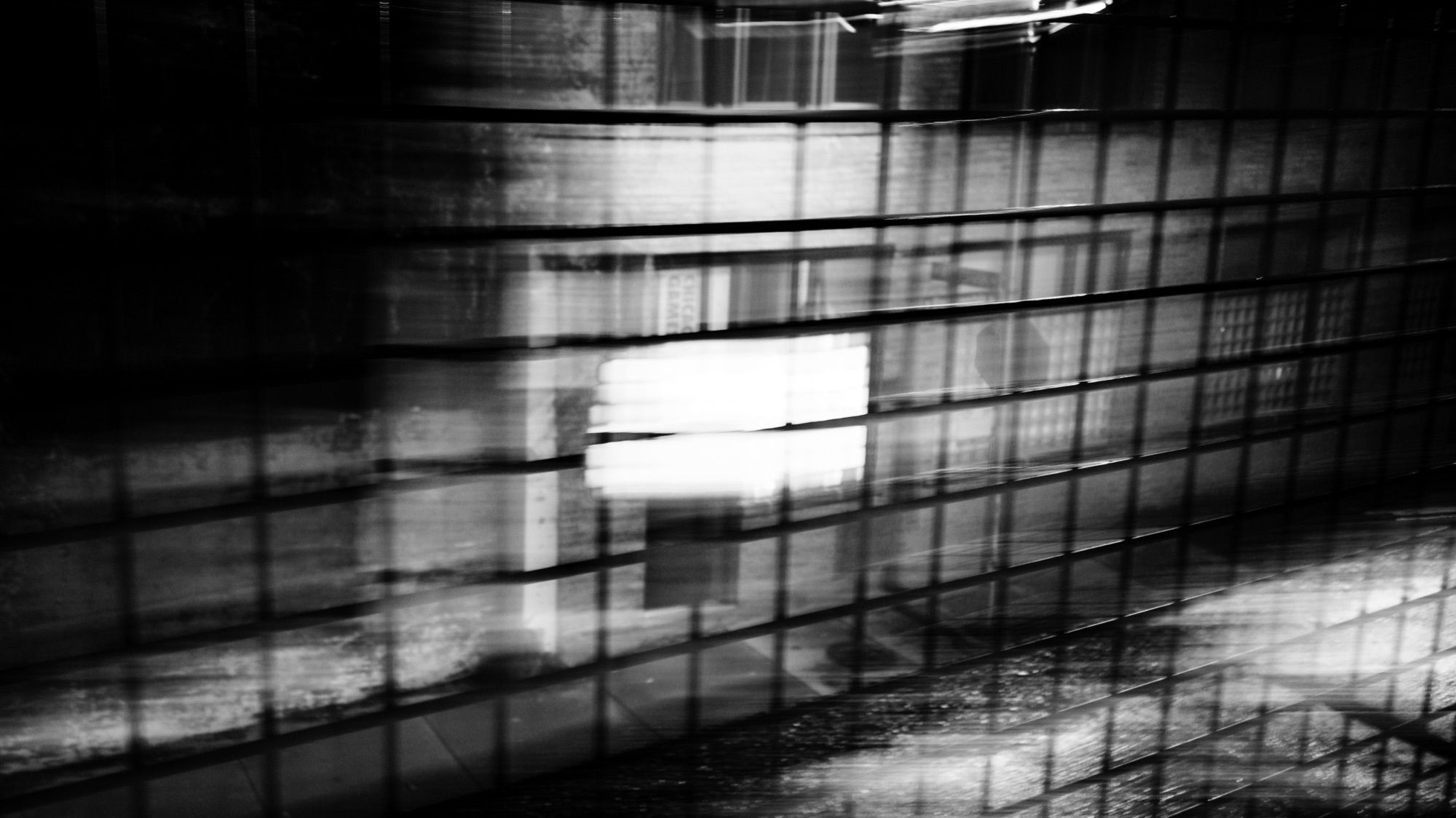 Nighttime. A scene of a side street/alley shot through fencing along the 606/Bloomingdale Trail. Extreme camera movement and a long shutter speed create stylistic blurring and light trails. Luminance contrast is high.