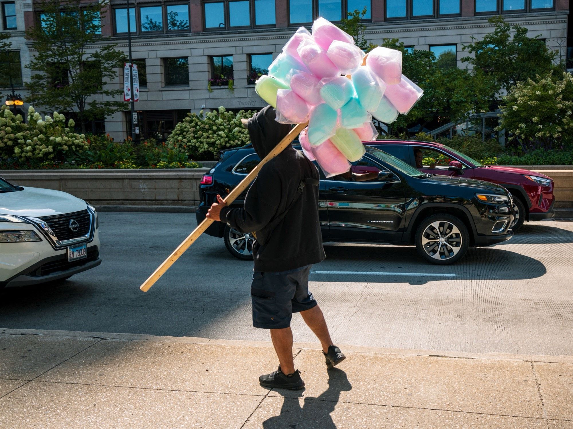 Daytime. A man holds a large stick across his shoulder, with bags of cotton candy attached at the higher end, on a sidewalk. Cars on Michigan Avenue are seen in his background. Colors are slightly stylized. Luminance contrast is mildly high.