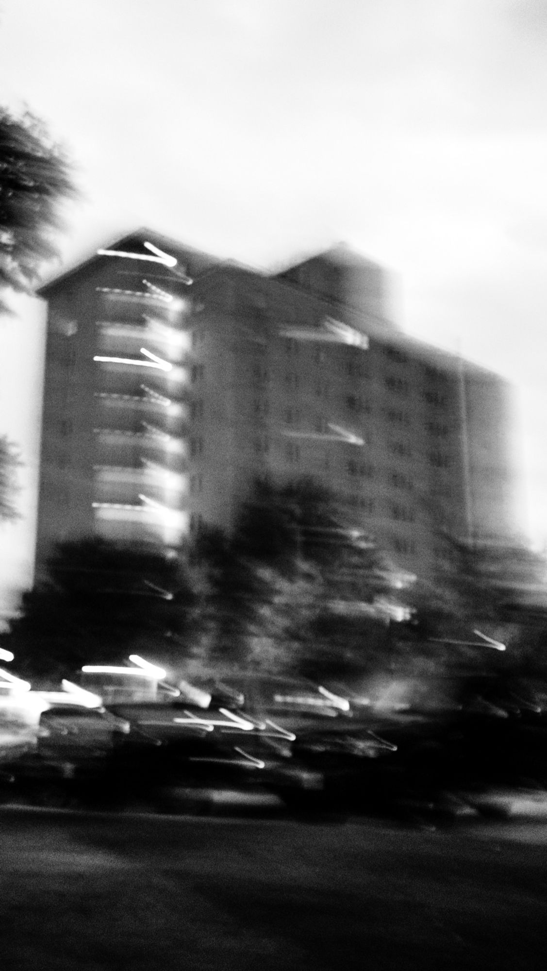 Nighttime. An abstraction of a large building set against they sky, with a car visible in the foreground. Branches of a tree can be seen in the left side of the frame. Extreme camera movement and a long shutter speed create blurring and light trails.