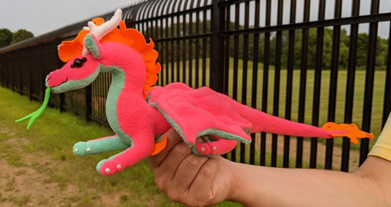 a bright neon pink dragon with an aqua blue underbelly/underwings/ears, black shiny eyes, an orange mane/crest/elbow crests/tailtip, white antlerlike horns and claws, and a green tongue