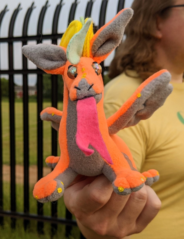 an orange dragon plushie, with a gray underbelly/underwings/inner ears, light blue mane/crest, elbow spines, and tailtip, yellow claws and semicircular horns, a pink tongue, and crystally eyes