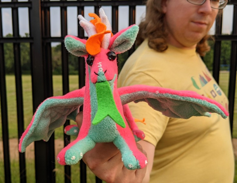 a bright neon pink dragon with an aqua blue underbelly/underwings/ears, black shiny eyes, an orange mane/crest/elbow crests/tailtip, white antlerlike horns and claws, and a green tongue