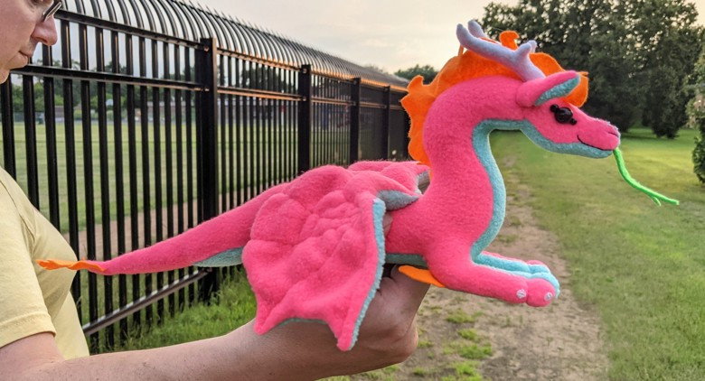 a bright neon pink dragon with an aqua blue underbelly/underwings/ears, black shiny eyes, an orange mane/crest/elbow crests/tailtip, white antlerlike horns and claws, and a green tongue